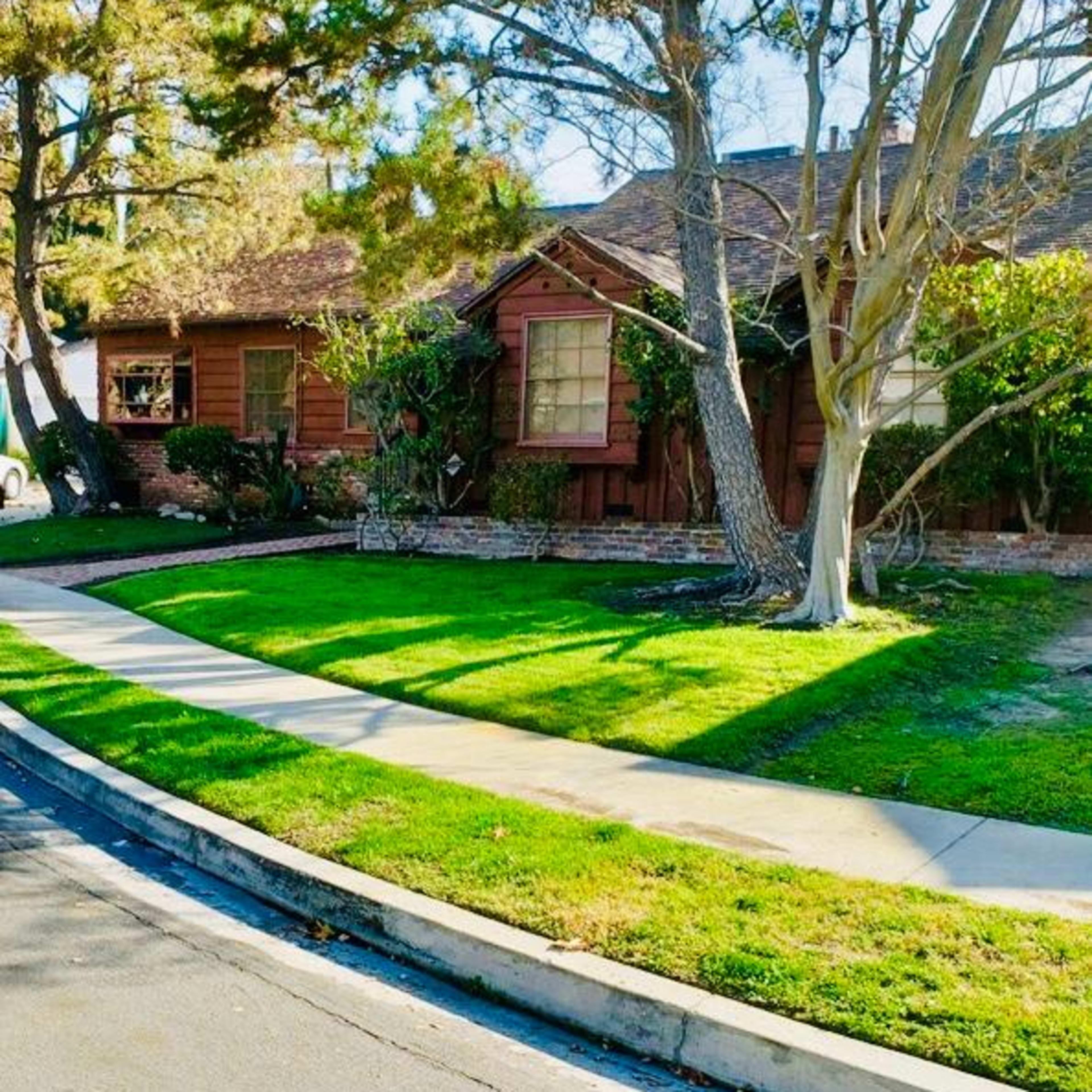 A brown wooden house with a pitched roof and several trees in the front yard is situated at the corner of a sidewalk and a street.