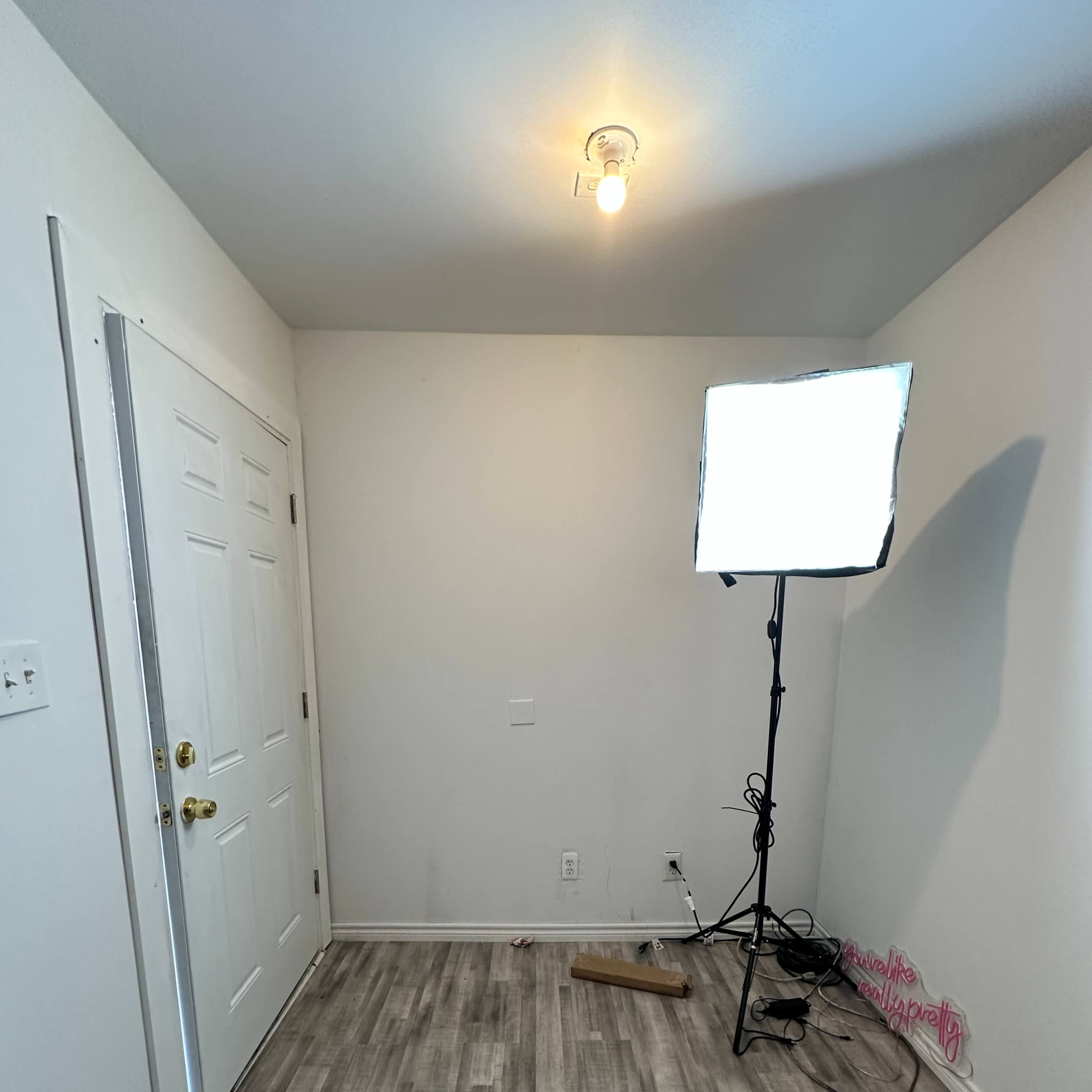 A small, empty room with a door on one wall, a light stand with a softbox, and gray wooden flooring.