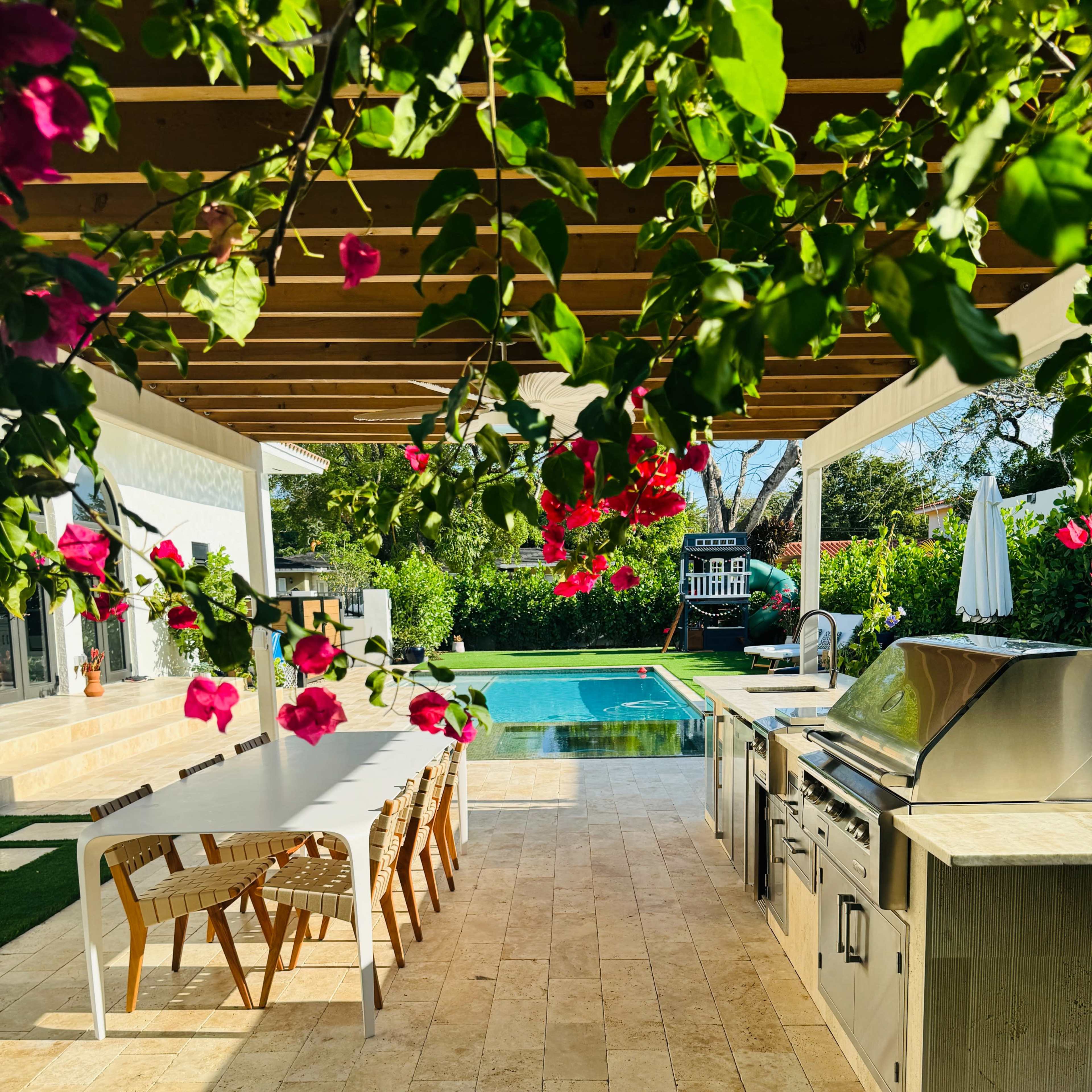 A modern outdoor kitchen and dining area with a view of a swimming pool, surrounded by lush greenery and blooming flowers.