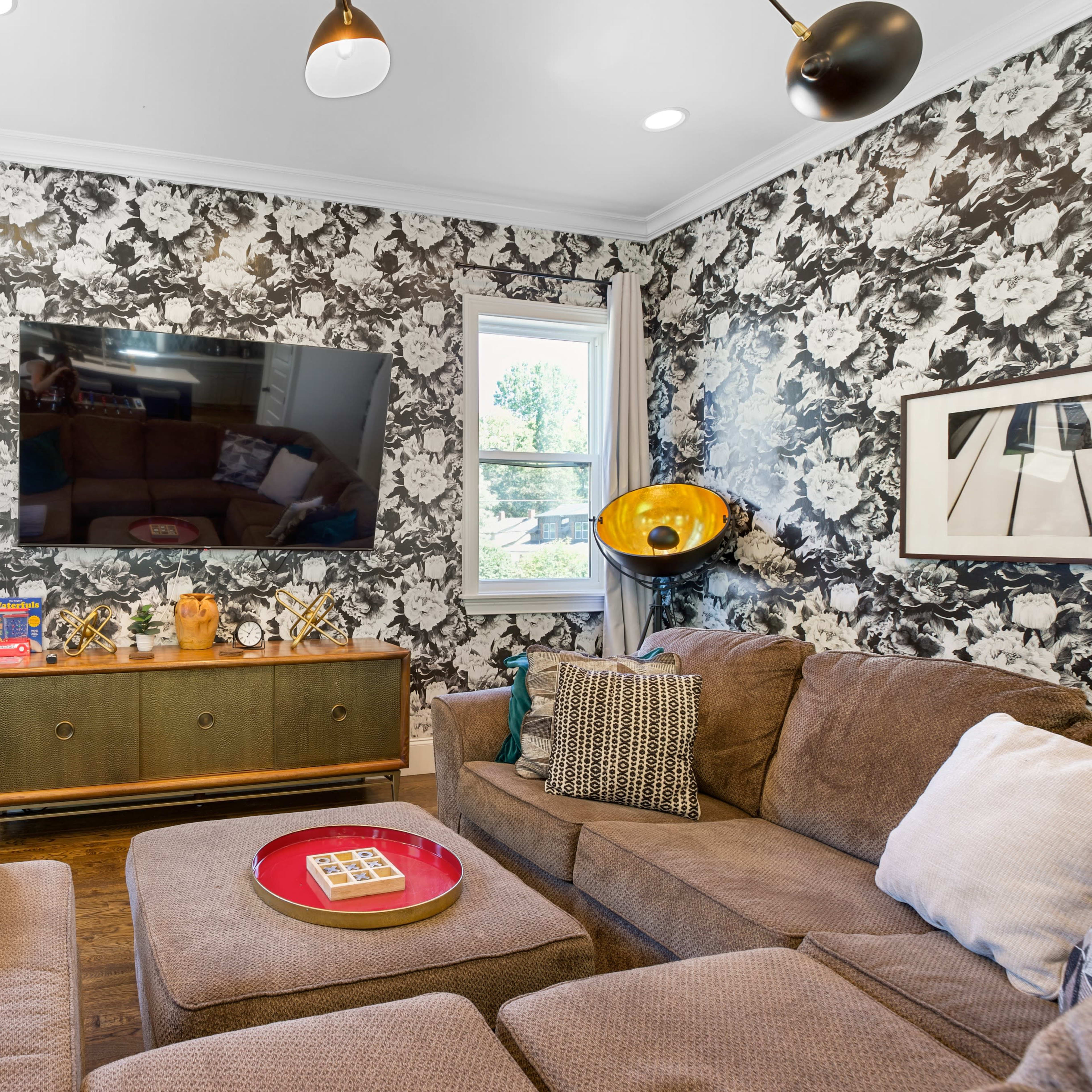 A living room featuring a textured floral wallpaper, a large television mounted on the wall, a brown sectional sofa, and various decorative items on a green sideboard.