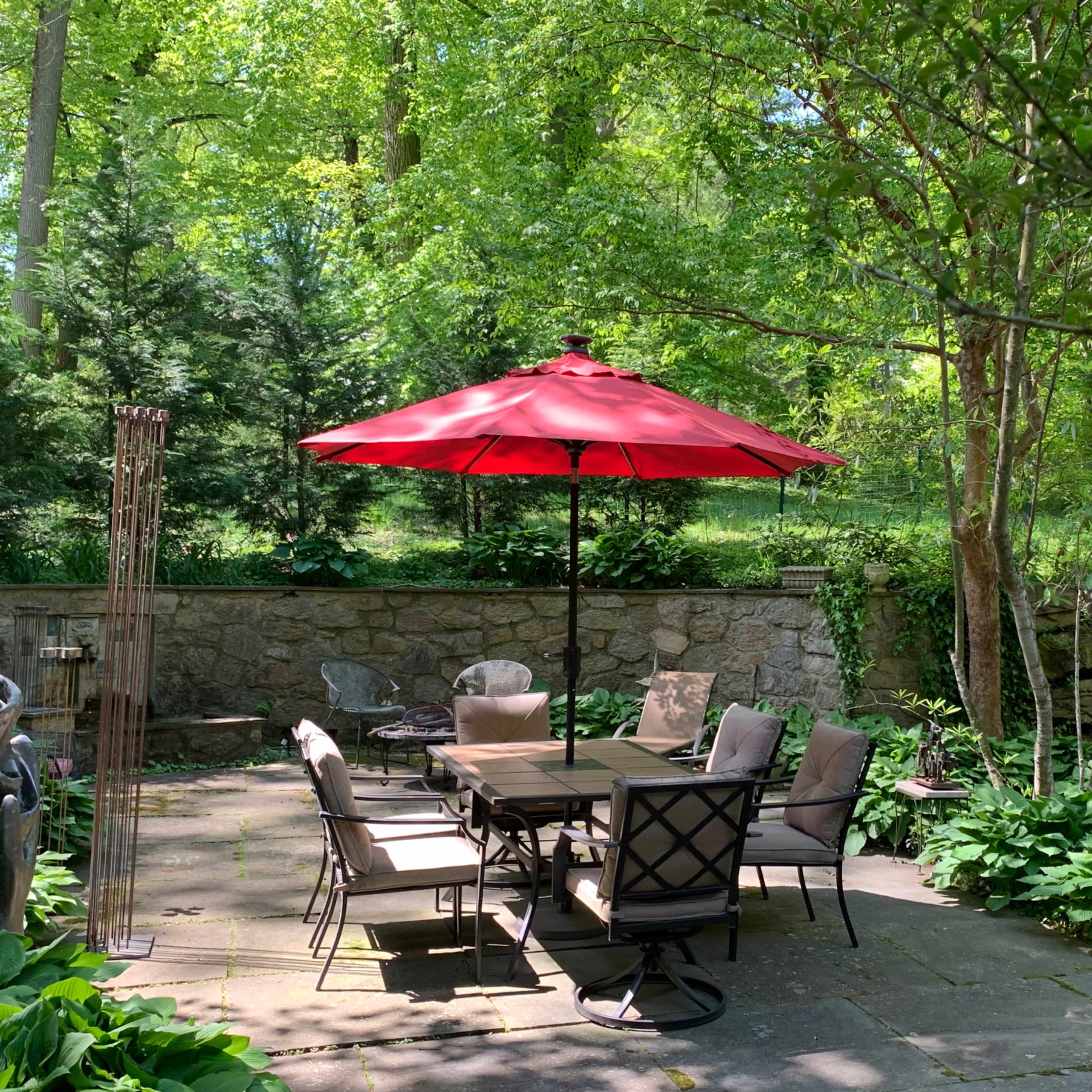 A shaded outdoor patio features a round table with chairs beneath a red umbrella, surrounded by greenery.