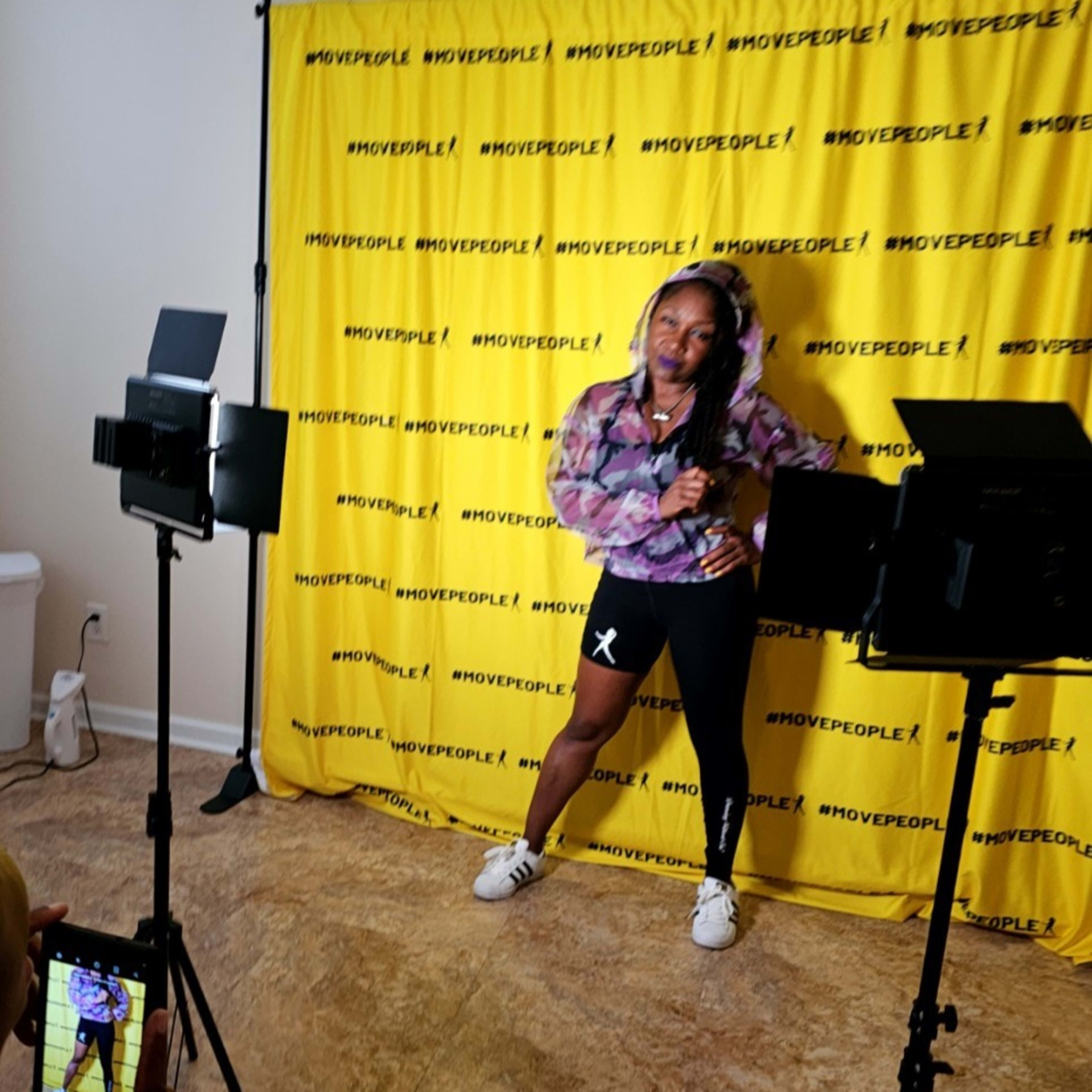 A person poses against a yellow backdrop with the text "#MOVEPEOPLE!" while two mounted lights illuminate the scene.