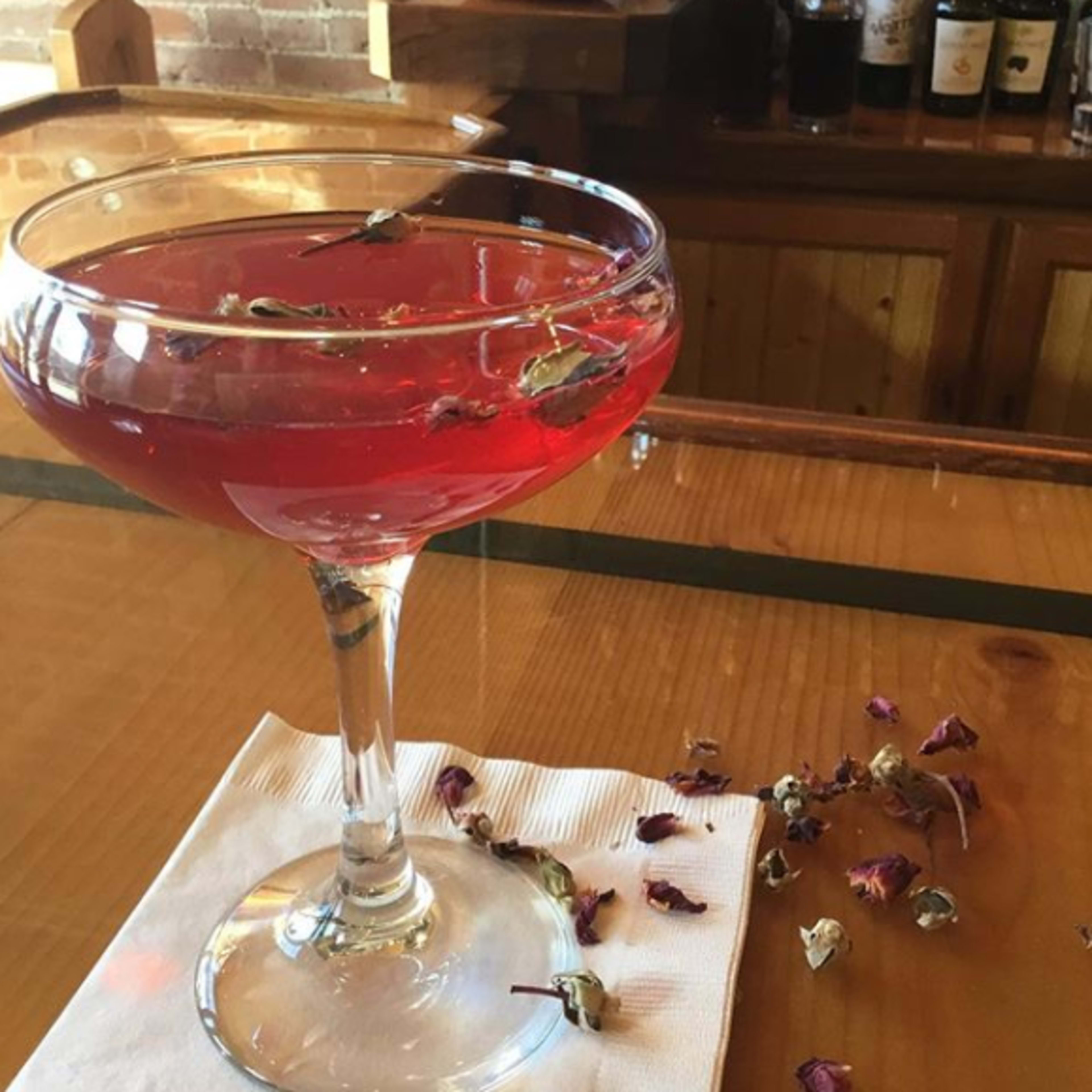 A cocktail glass filled with a red drink and garnished with dried rose petals rests on a wooden bar top beside scattered flower petals.