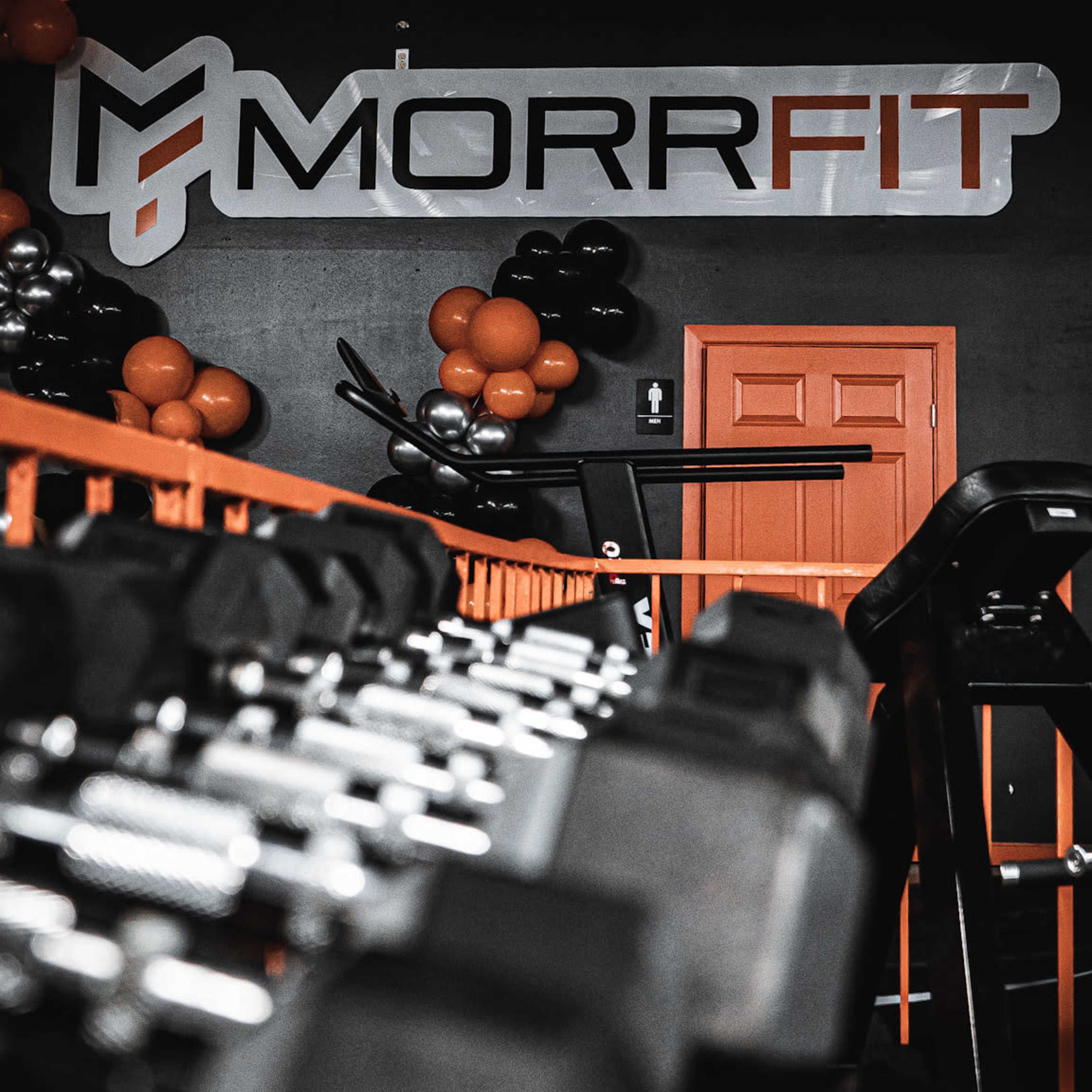 A gym interior featuring a wall with the "MORRFIT" logo and a rack of dumbbells in the foreground.