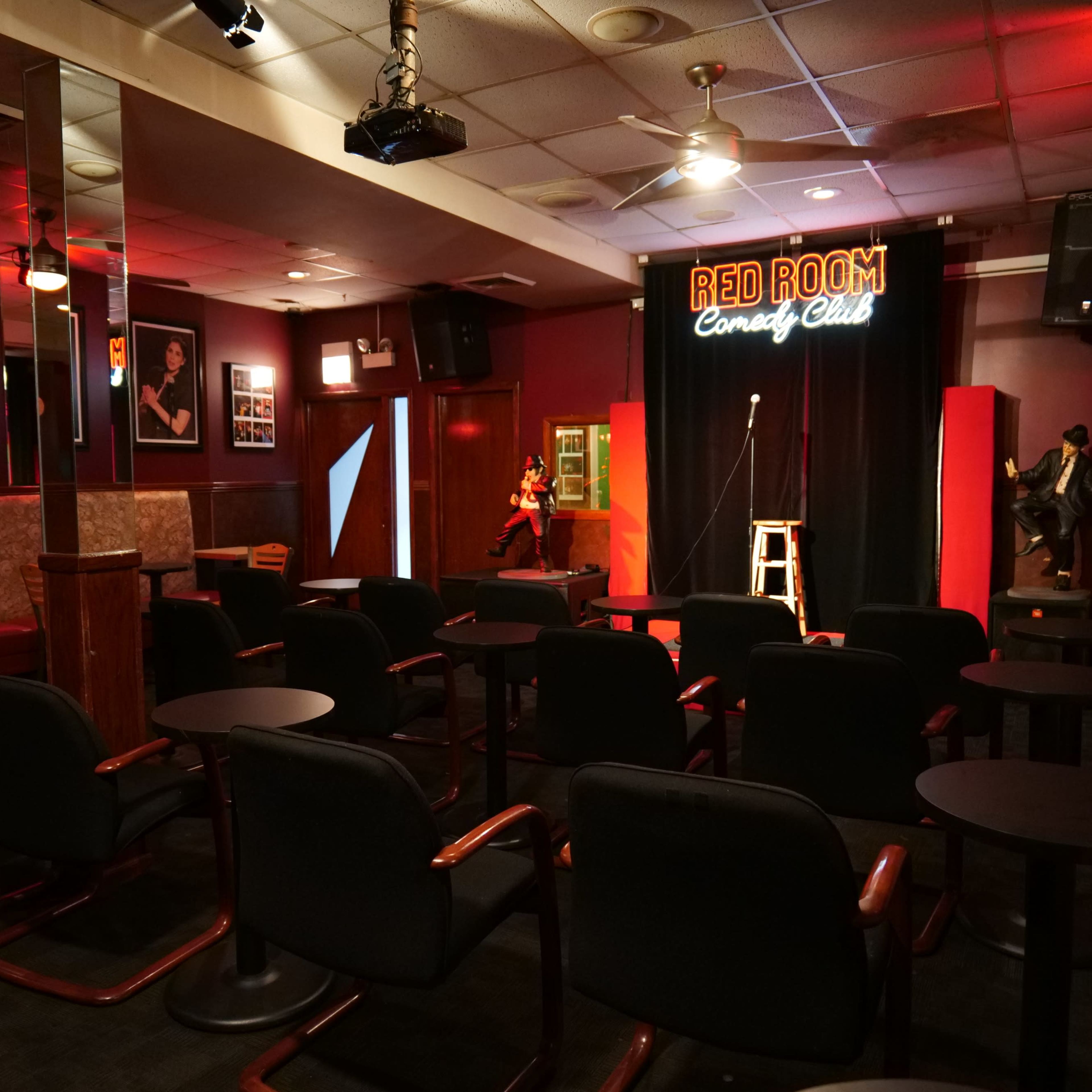 A comedy club with empty chairs arranged in front of a stage featuring a microphone and a stool, illuminated by a sign that reads "RED ROOM Comedy Club."