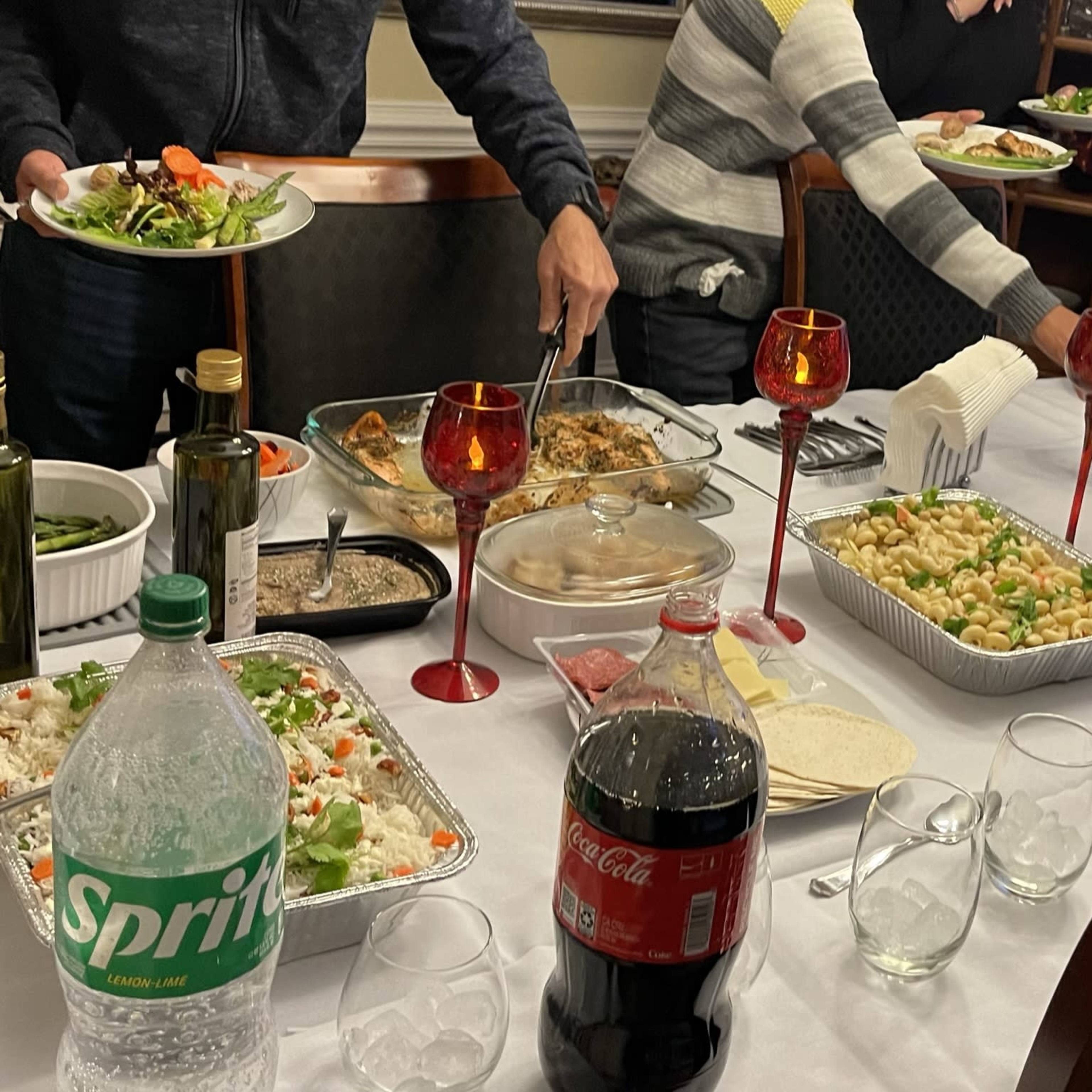 A dining table is set with various food dishes, drinks, and people serving themselves.