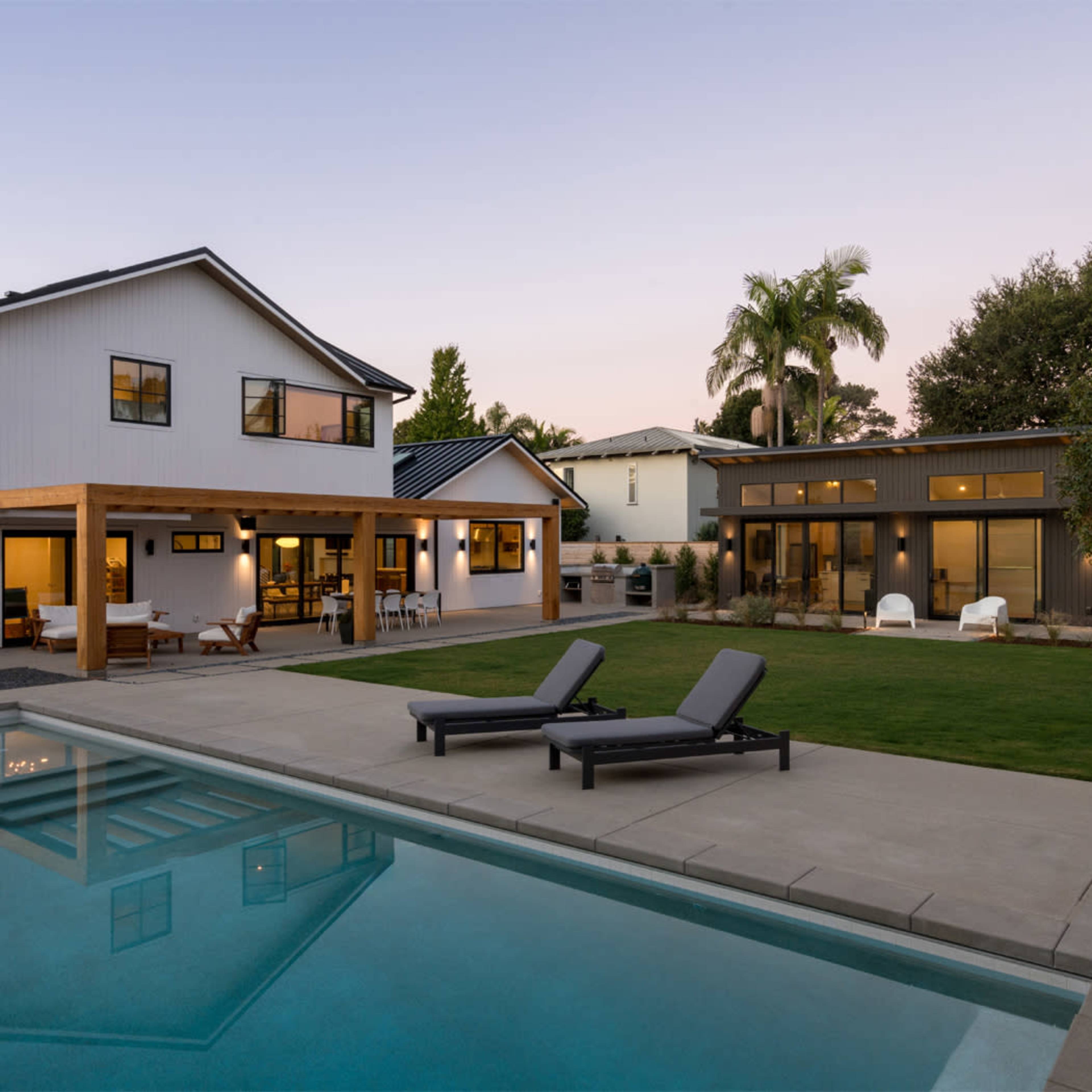 A modern backyard features a pool with lounge chairs, adjacent to two contemporary-style houses surrounded by greenery at dusk.