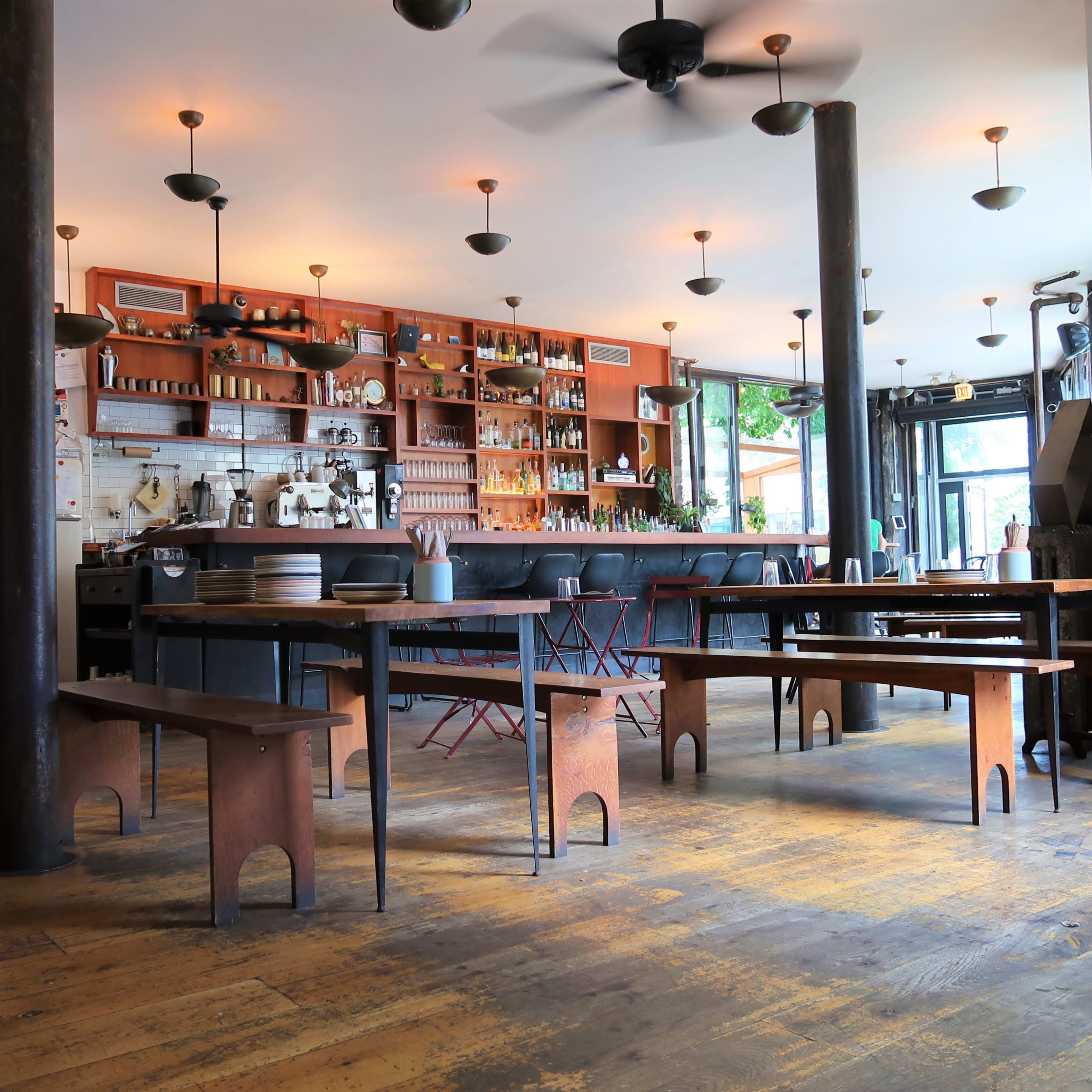 A spacious café interior with wooden flooring, a bar area with a variety of bottles, and several tables arranged throughout the room.