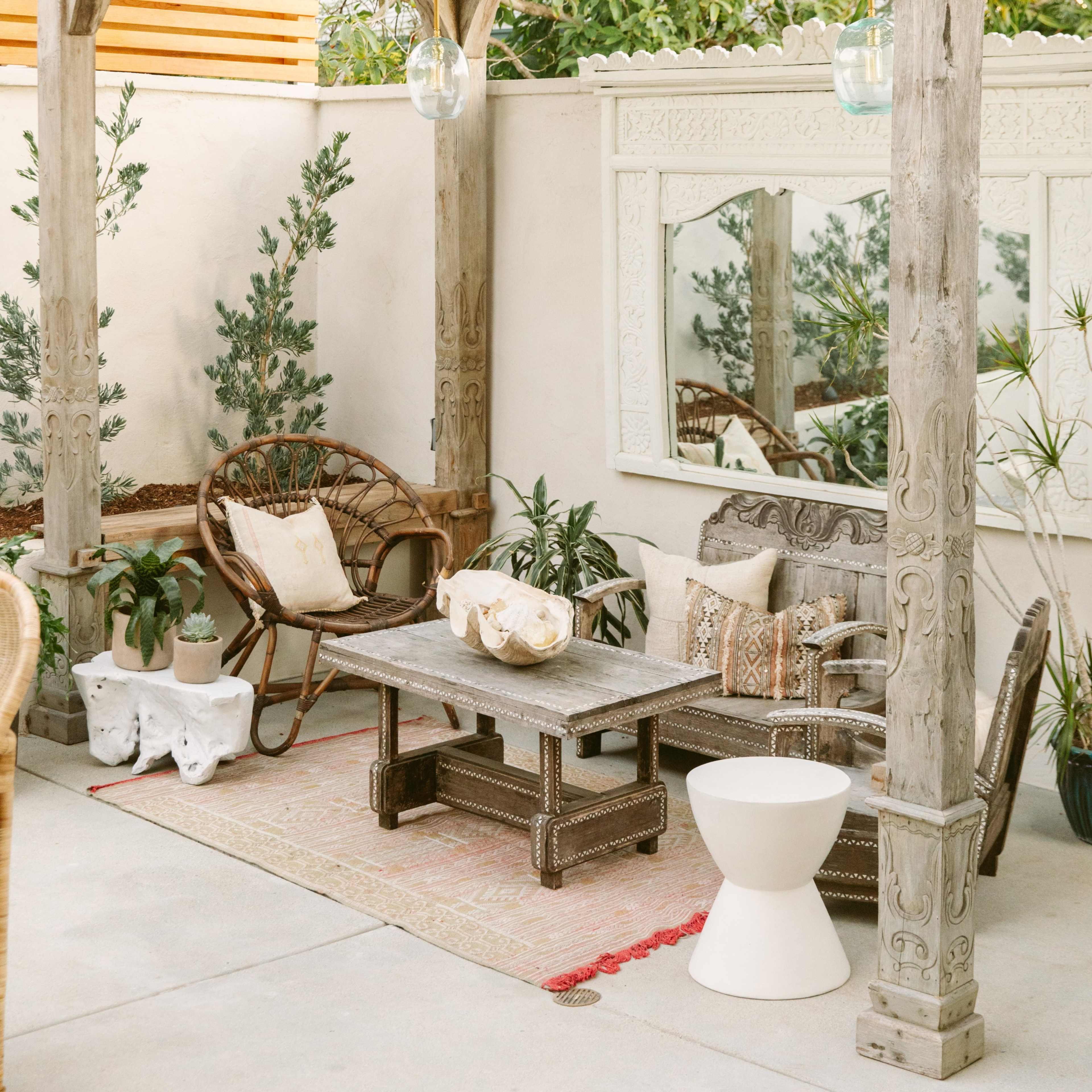 A cozy outdoor seating area featuring a wooden pergola with decorative details, a low table in the center, and various plants arranged around the space.
