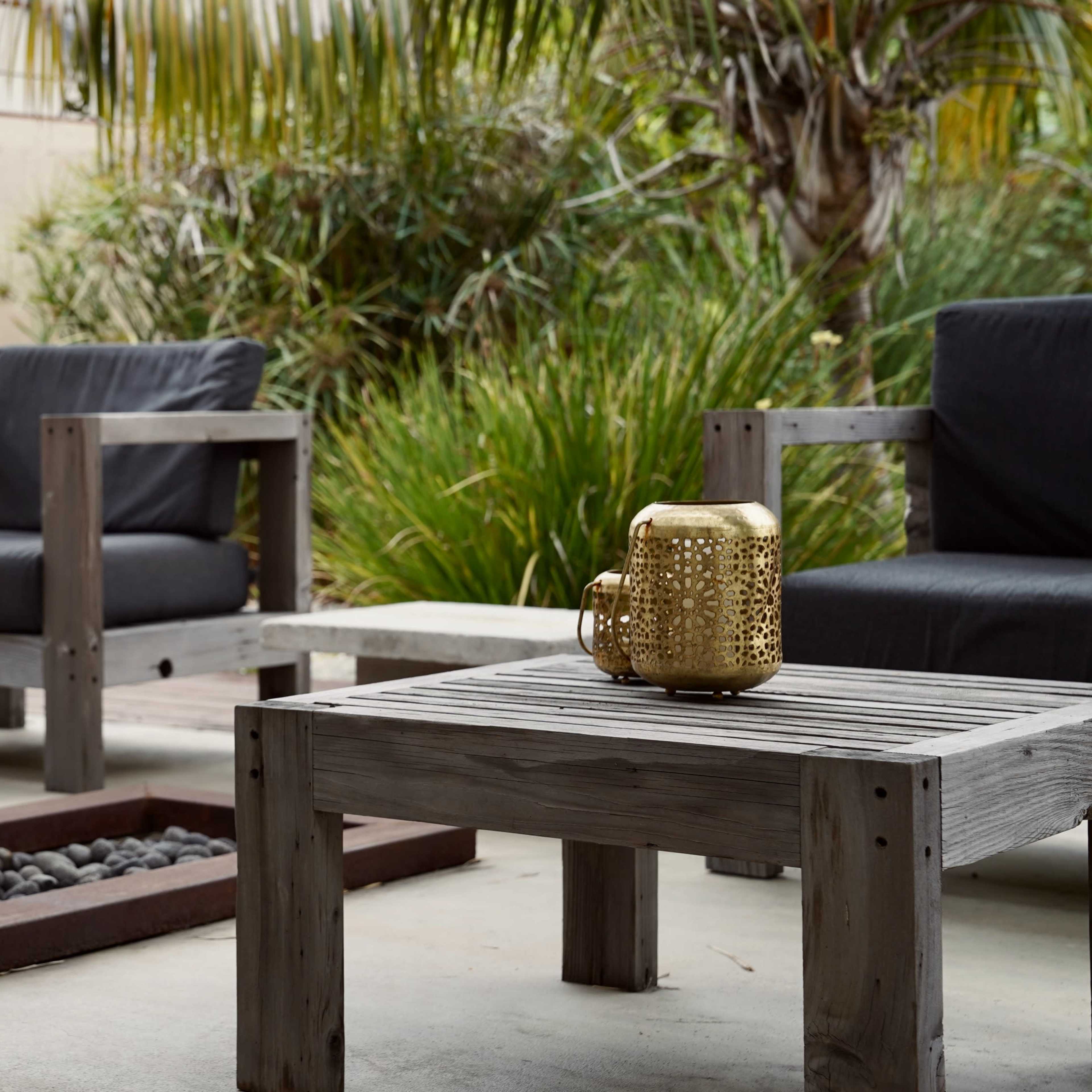 A patio area with wooden furniture, a stone fire pit filled with rocks, and greenery in the background.