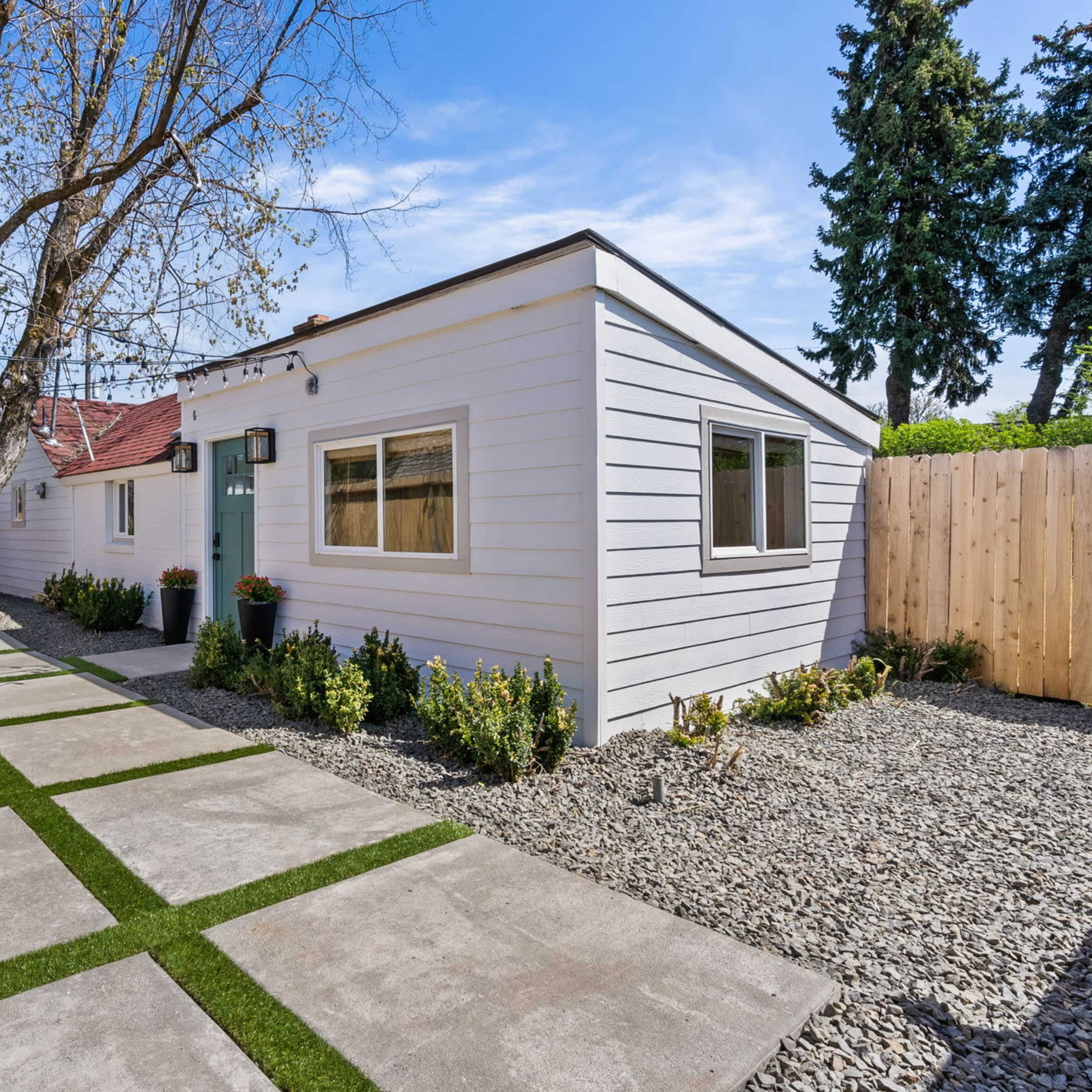 A narrow pathway lined with gravel and grass leads to a light-colored house surrounded by a wooden fence and a large tree.