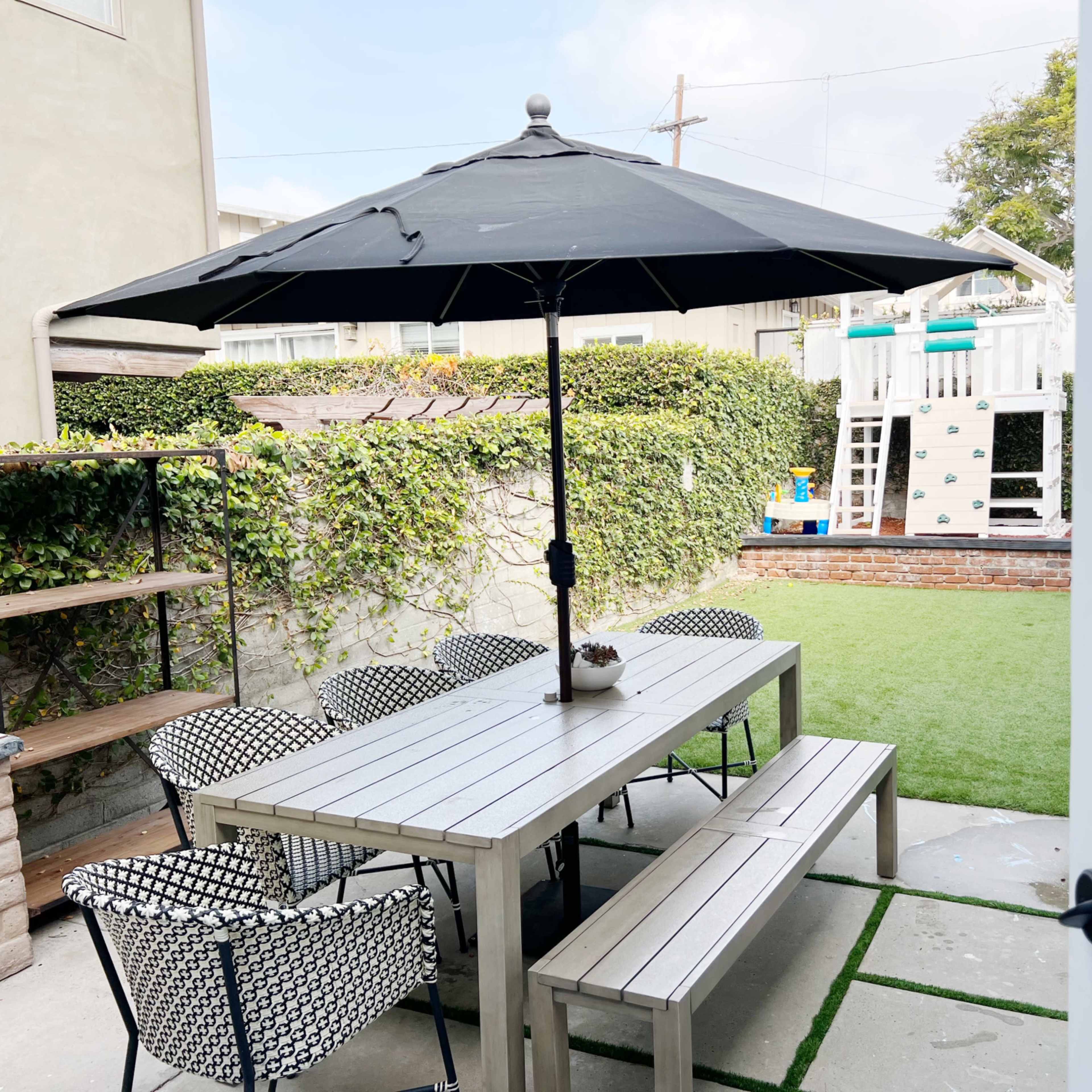 A patio area features a large table with chairs under an umbrella, surrounded by greenery and a playset in the background.