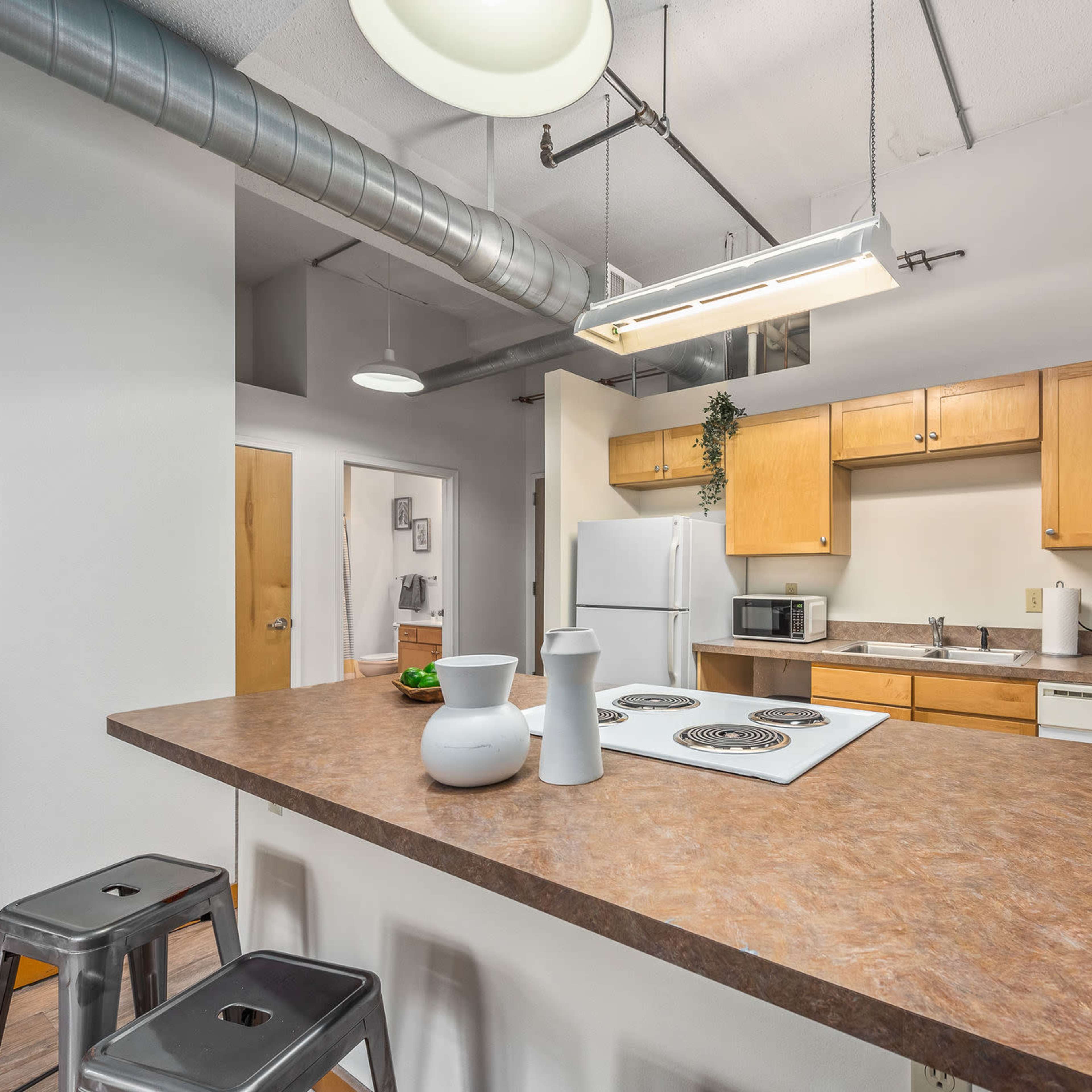 A modern kitchen in an apartment, featuring wooden cabinets, a white refrigerator, and a breakfast bar with two metal stools.