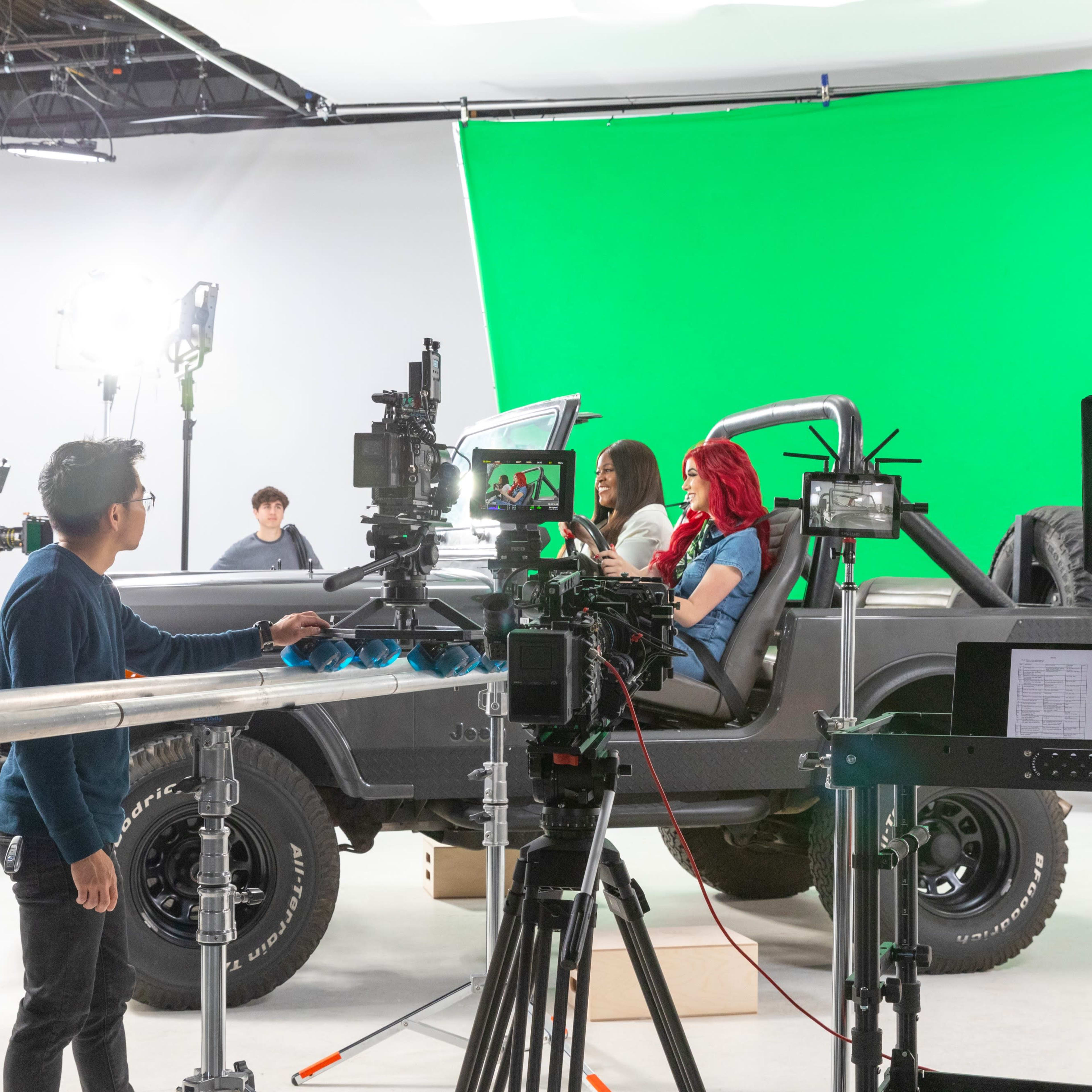A film crew operates cameras and lighting equipment around a vehicle on a soundstage with a green screen backdrop.