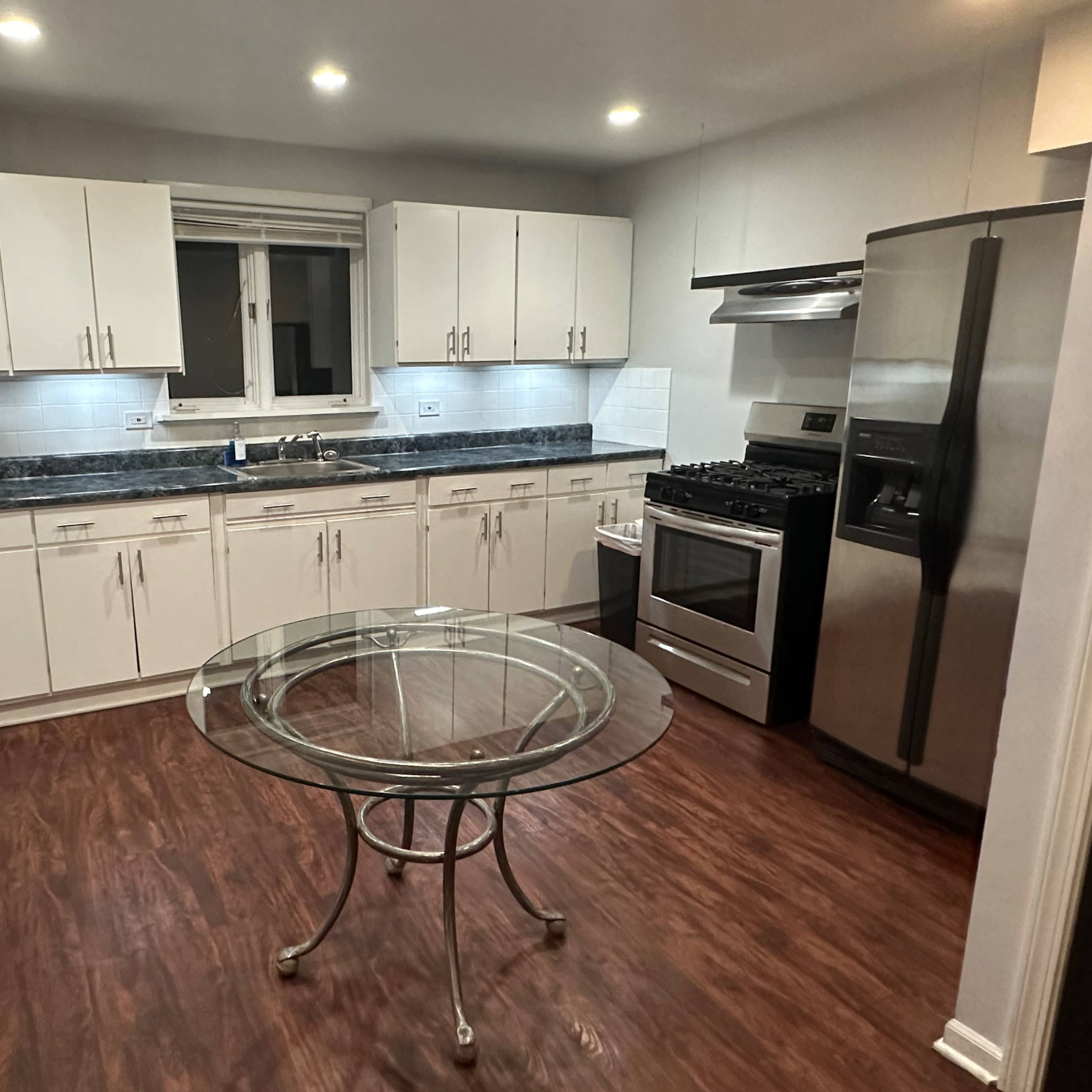 A modern kitchen with white cabinets, a stainless steel refrigerator, a gas stove, and a round glass dining table at the center.