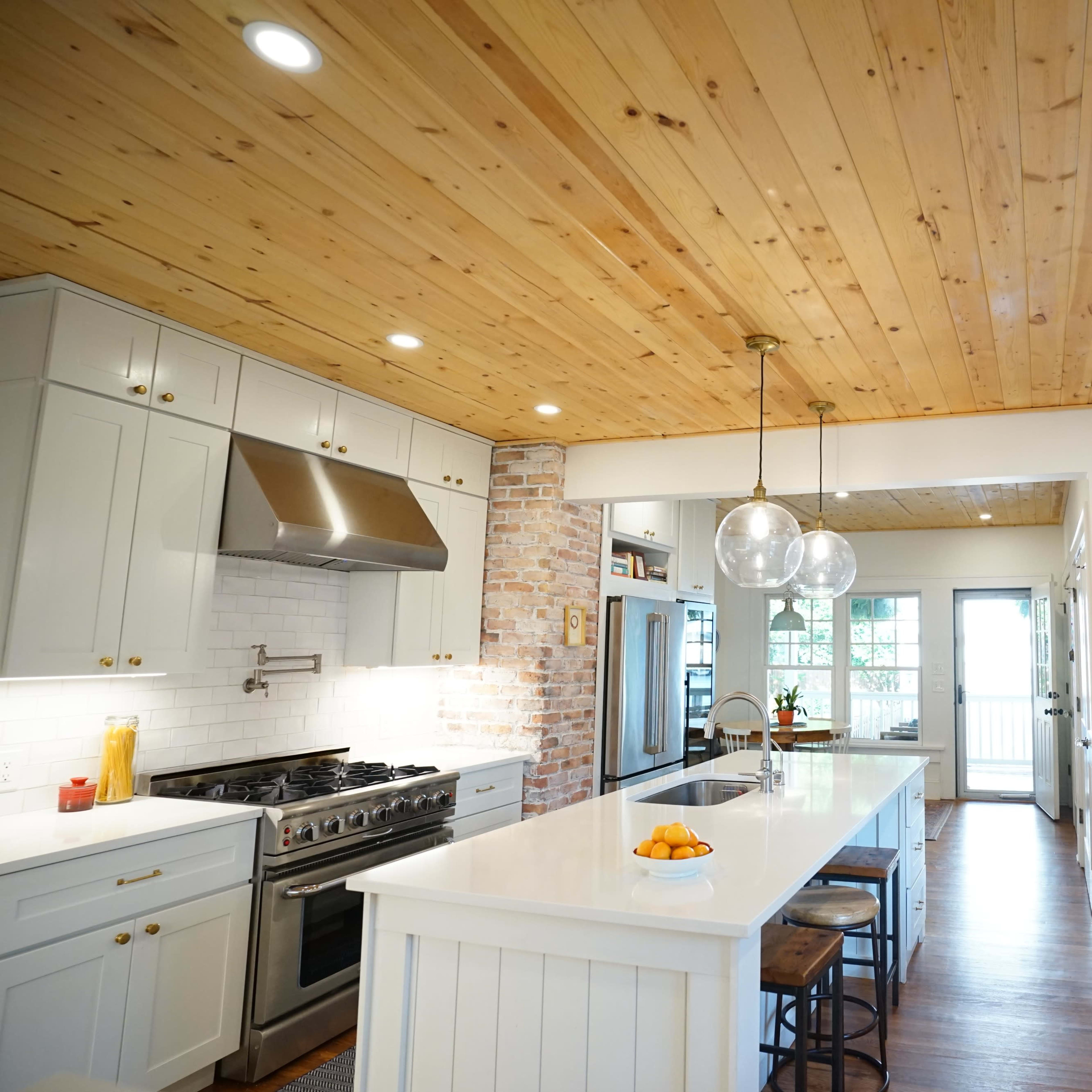 A bright kitchen features a large island with a white countertop, stainless steel appliances, and wooden accents including a ceiling and countertop.