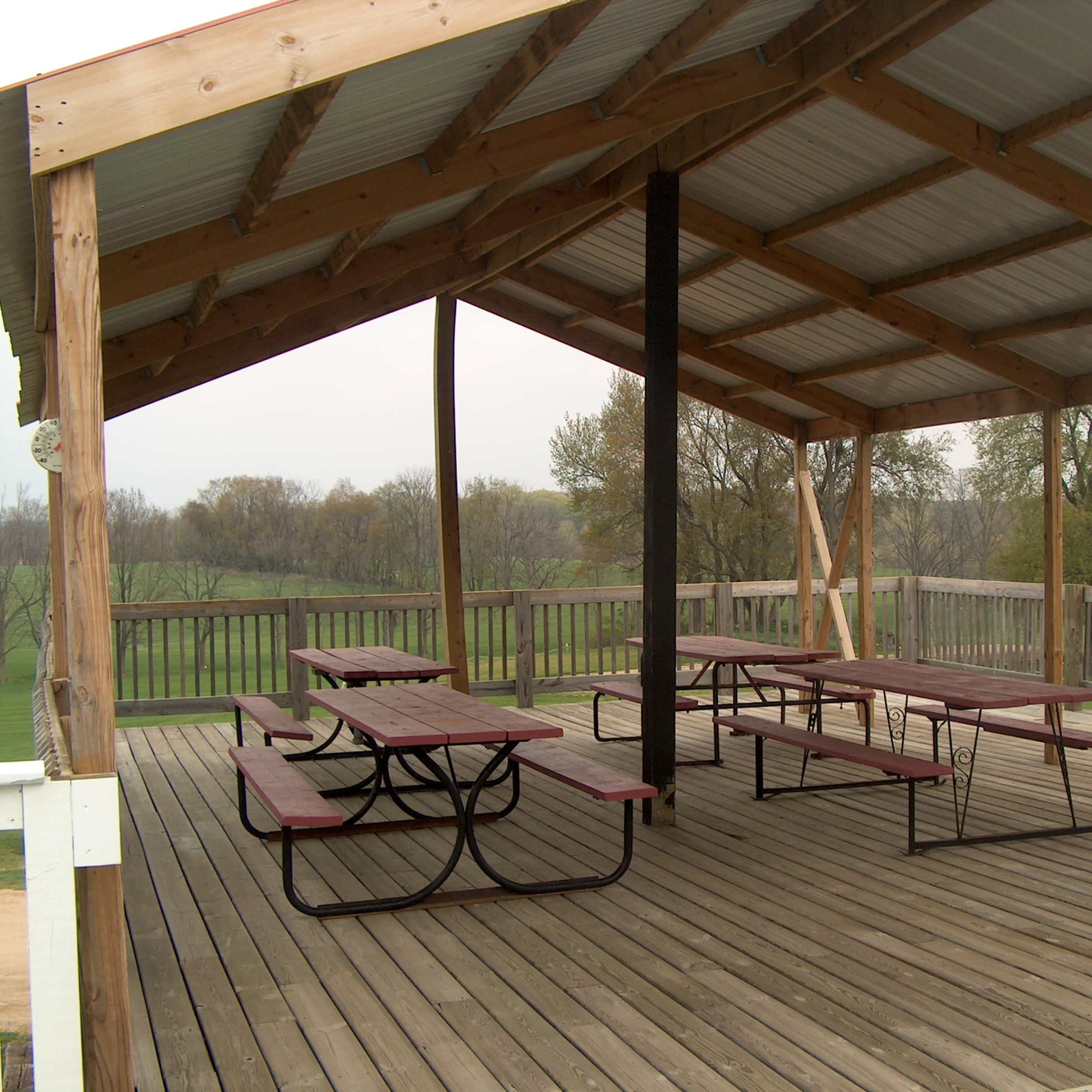 A sheltered structure with wooden decking and several picnic tables underneath, overlooking a grassy landscape.