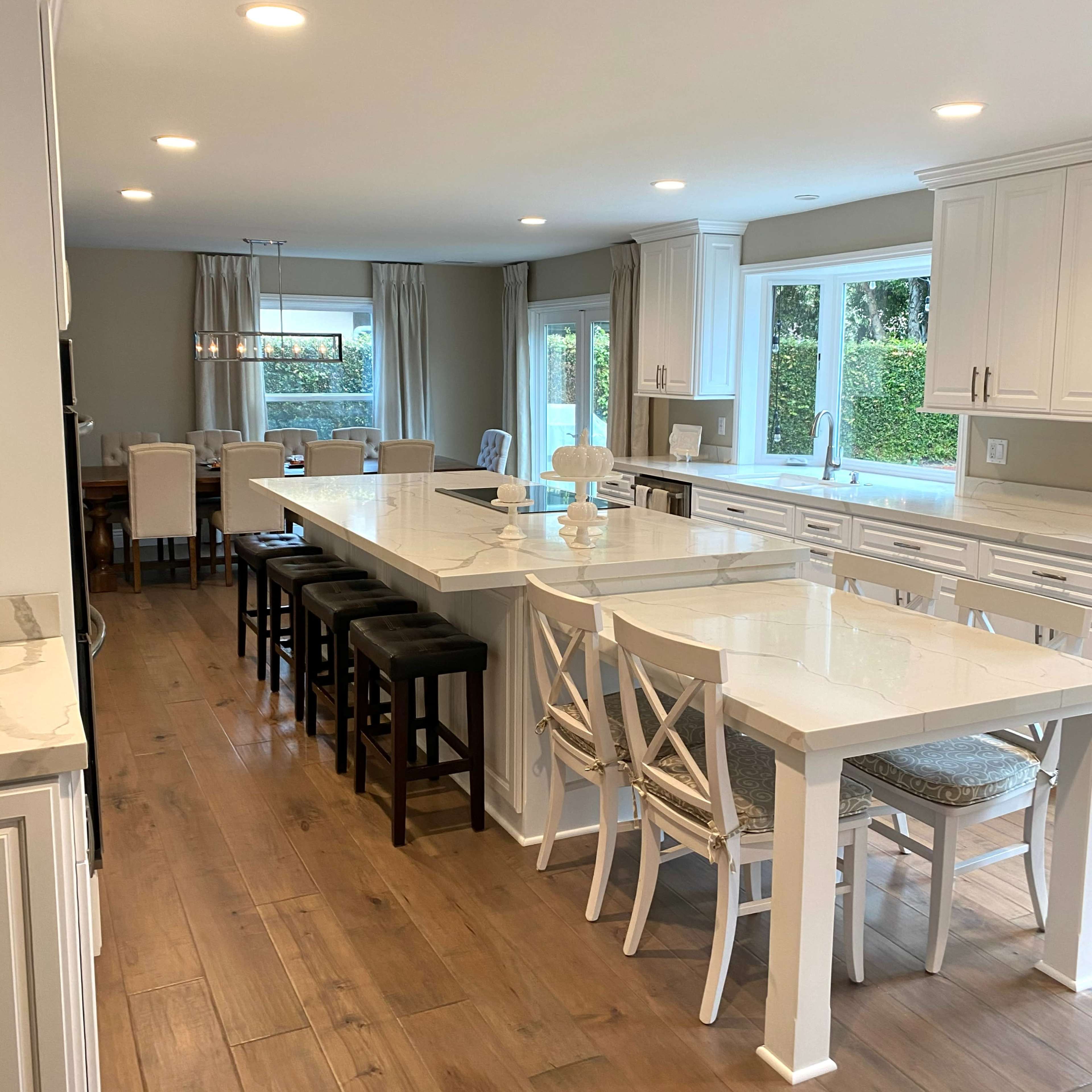 A modern kitchen features a large central island with bar stools, white cabinetry, and windows offering natural light.