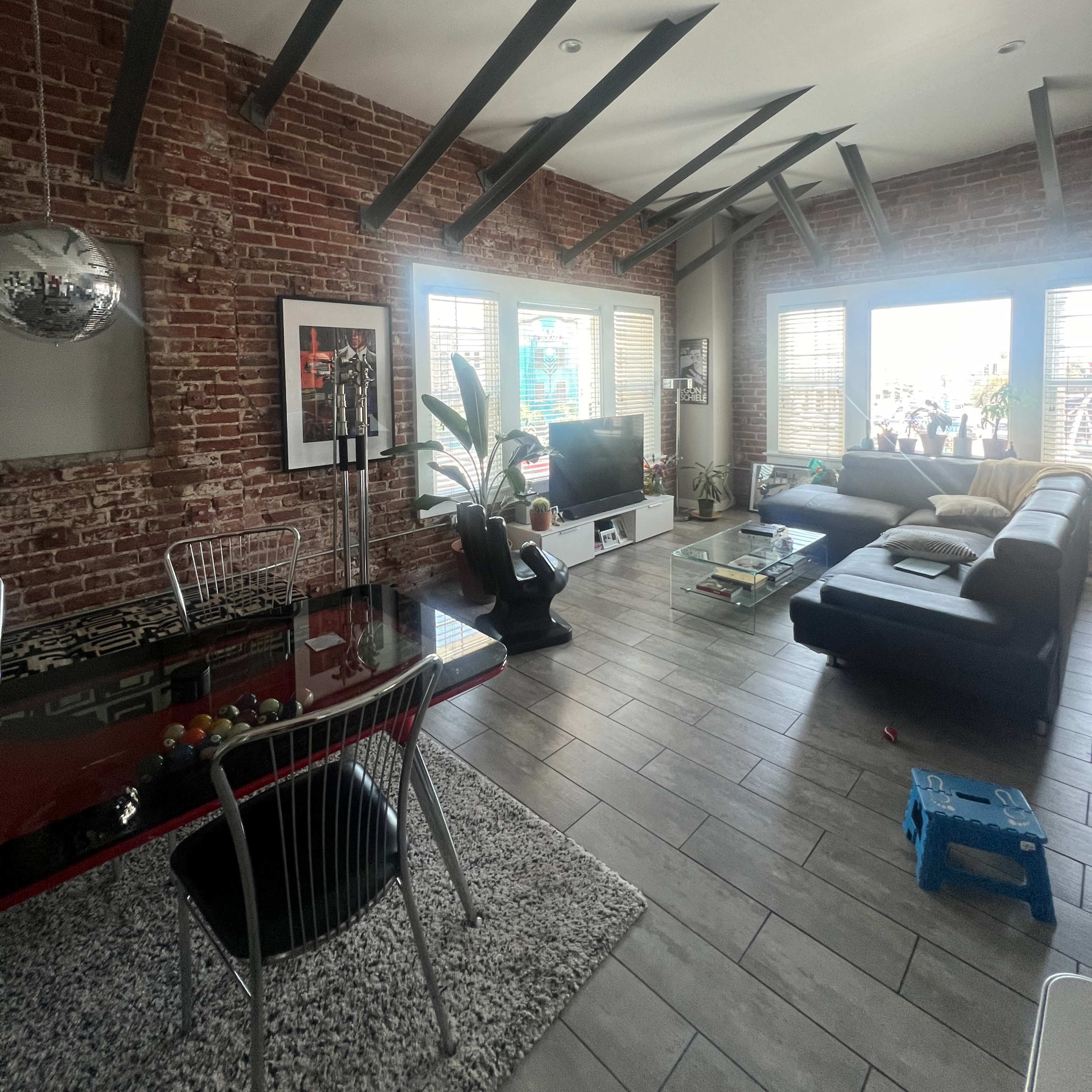 A modern living space with exposed brick walls, large windows letting in natural light, and a mix of contemporary furniture including a sectional sofa and a dining area with a pool table.
