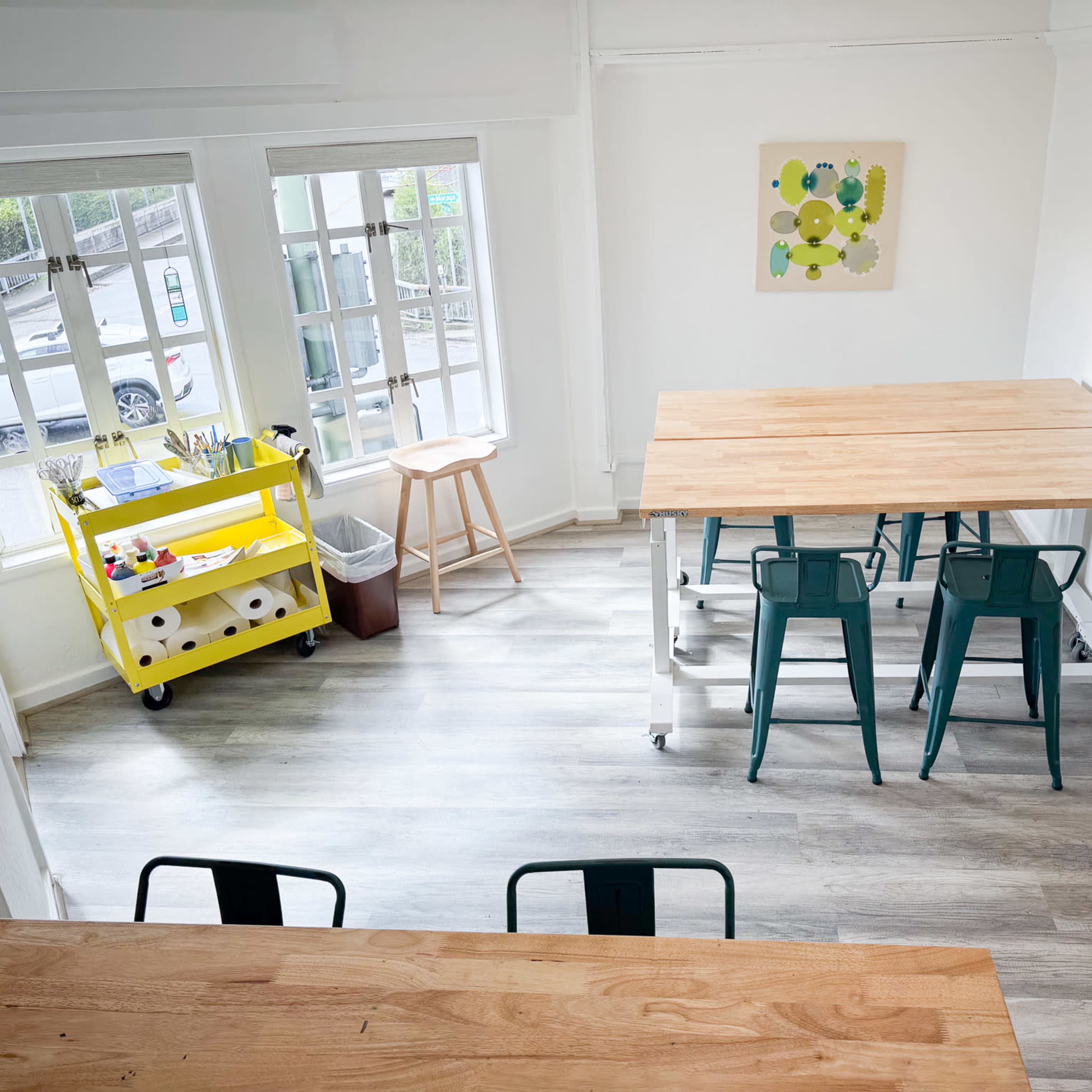 A bright, modern workspace featuring two tables, green chairs, a rolling cart with supplies, and large windows allowing natural light.