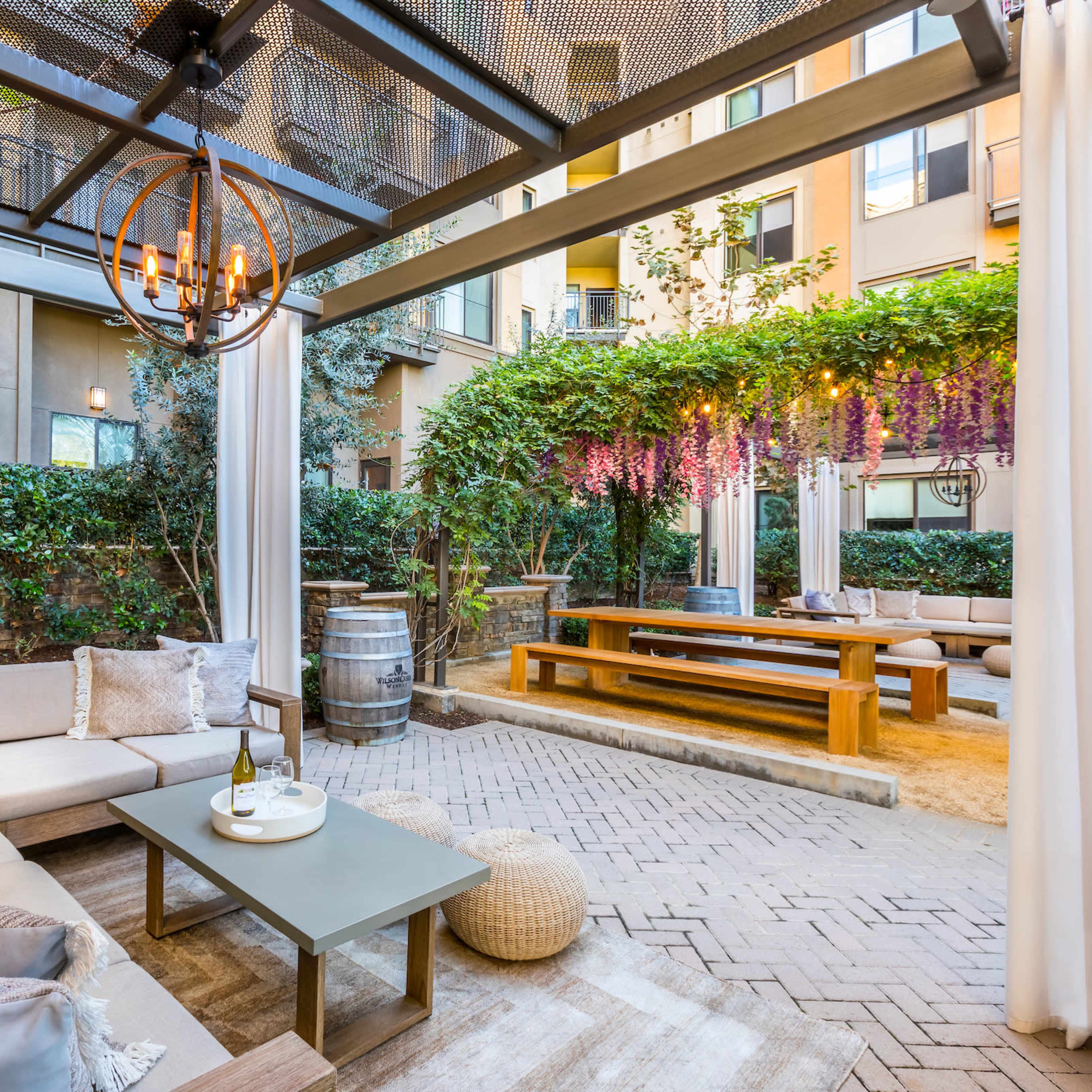 A landscaped outdoor seating area with a sofa, a coffee table, and a dining table surrounded by greenery and decorative lighting.