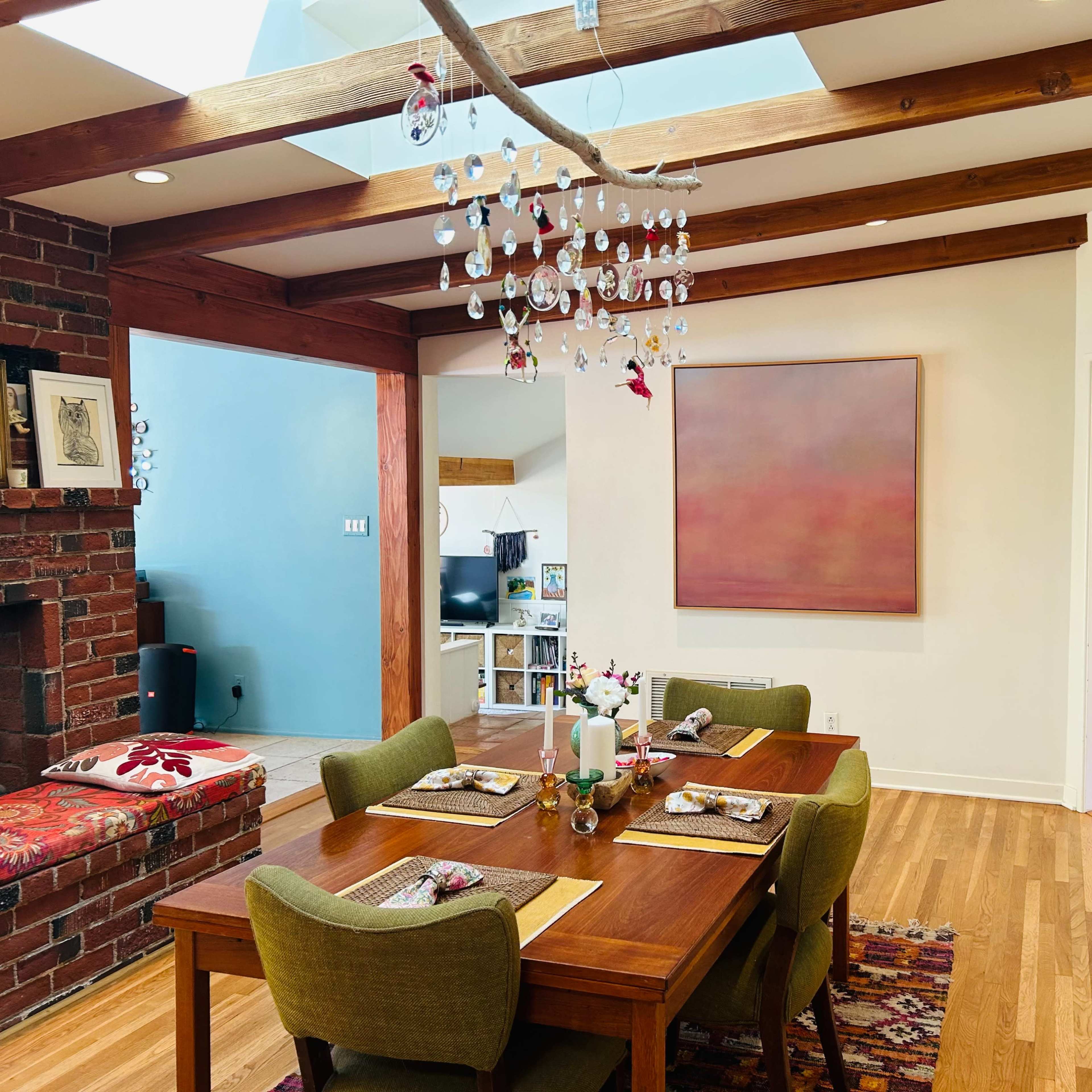 A dining area features a wooden table set for a meal, surrounded by green chairs, with a chandelier hanging above and artwork on the walls.