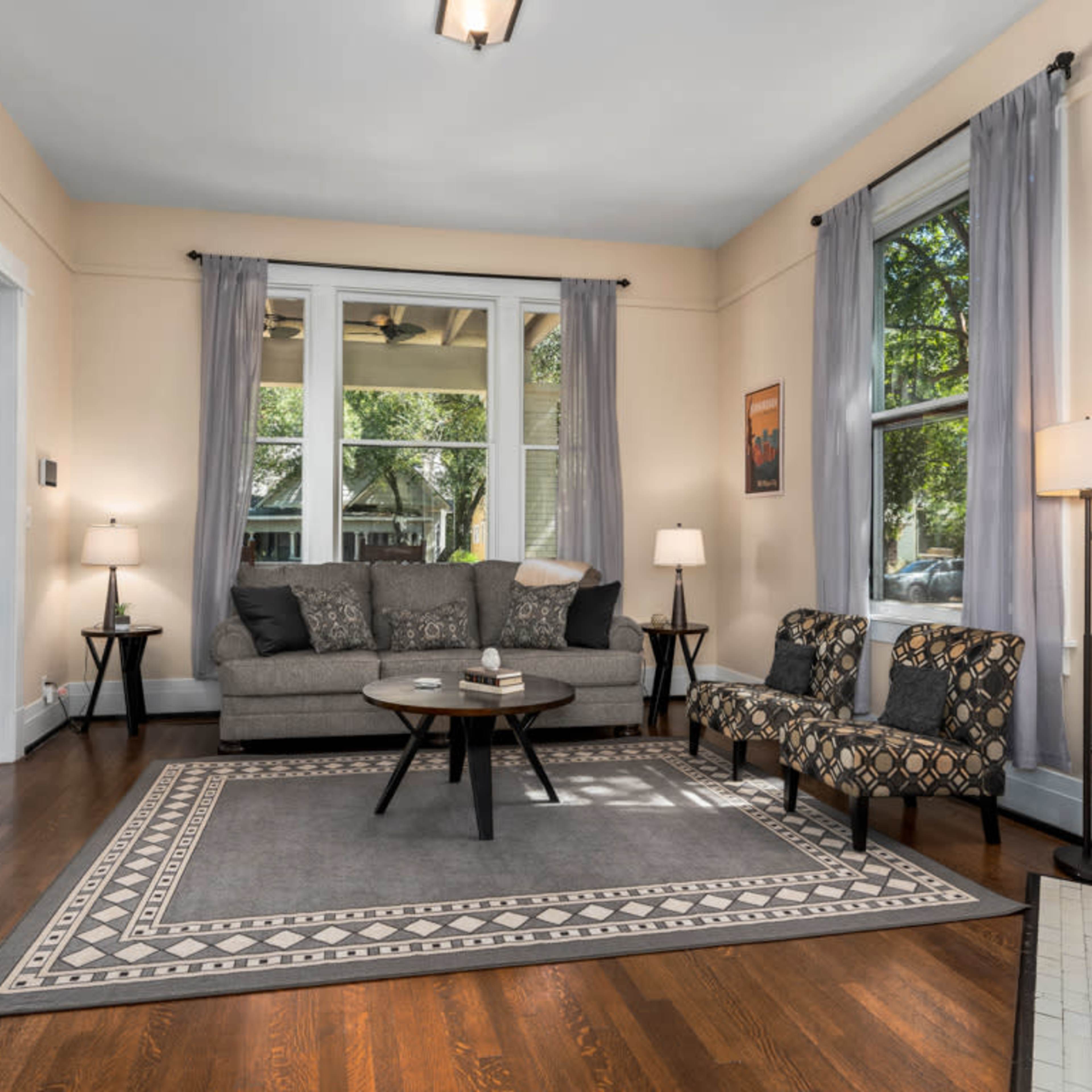 A living room with a gray sofa, a patterned chair, a coffee table, and large windows allowing natural light.