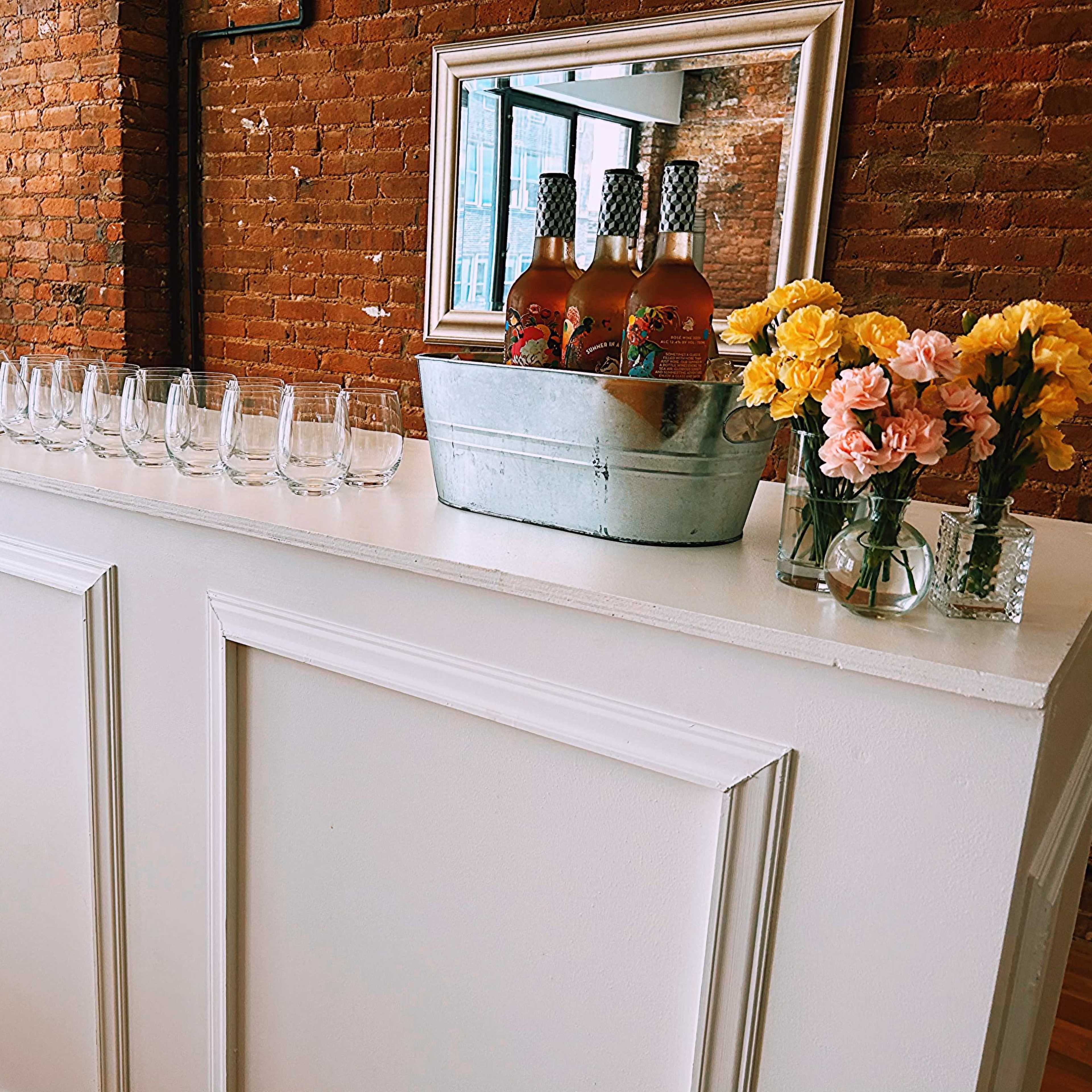 A white bar counter with a mirror above it displays several glasses and a metal bucket holding bottles, surrounded by colorful flower arrangements.