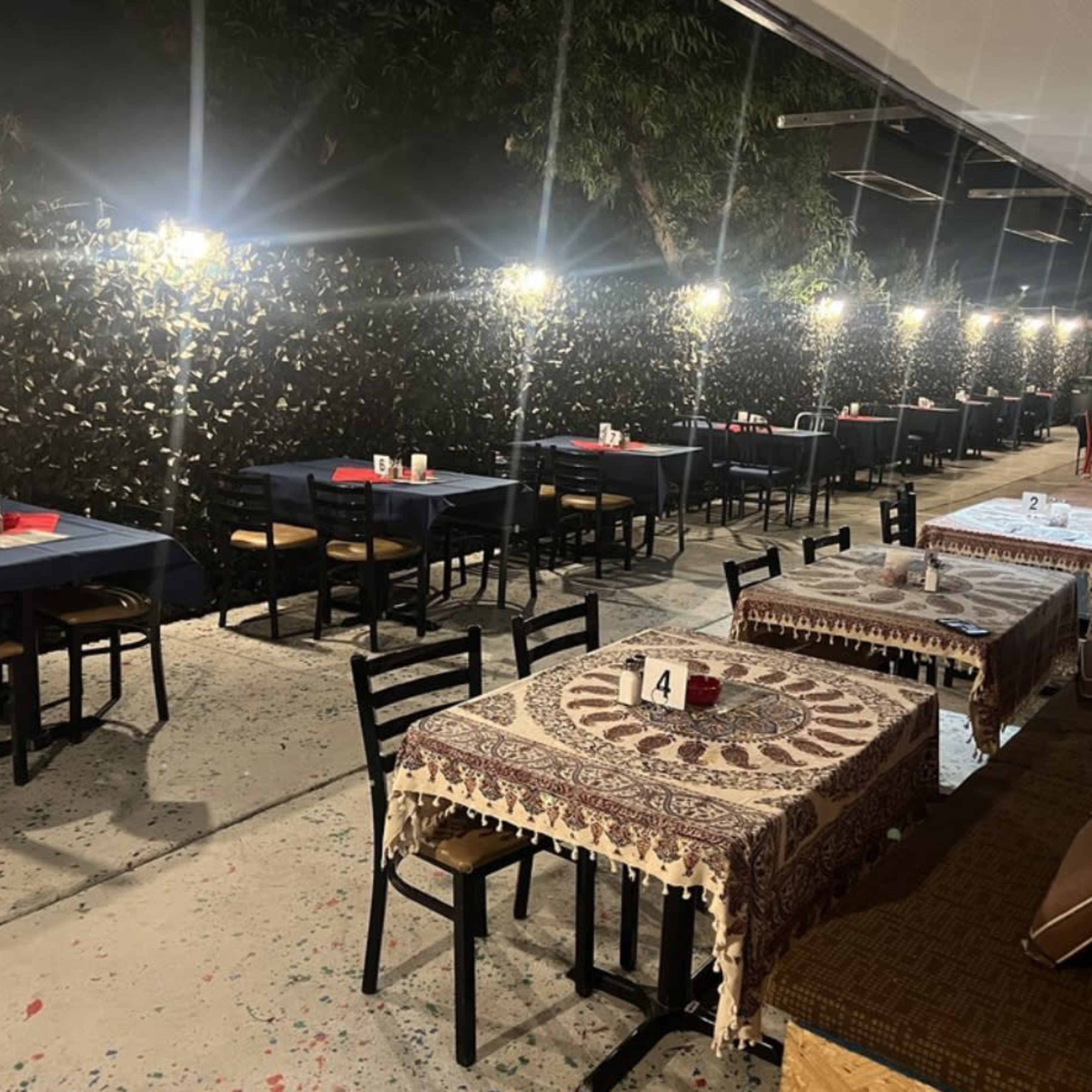 A dimly lit outdoor dining area with several tables set for customers, featuring tables covered with tablecloths and decorative runners.