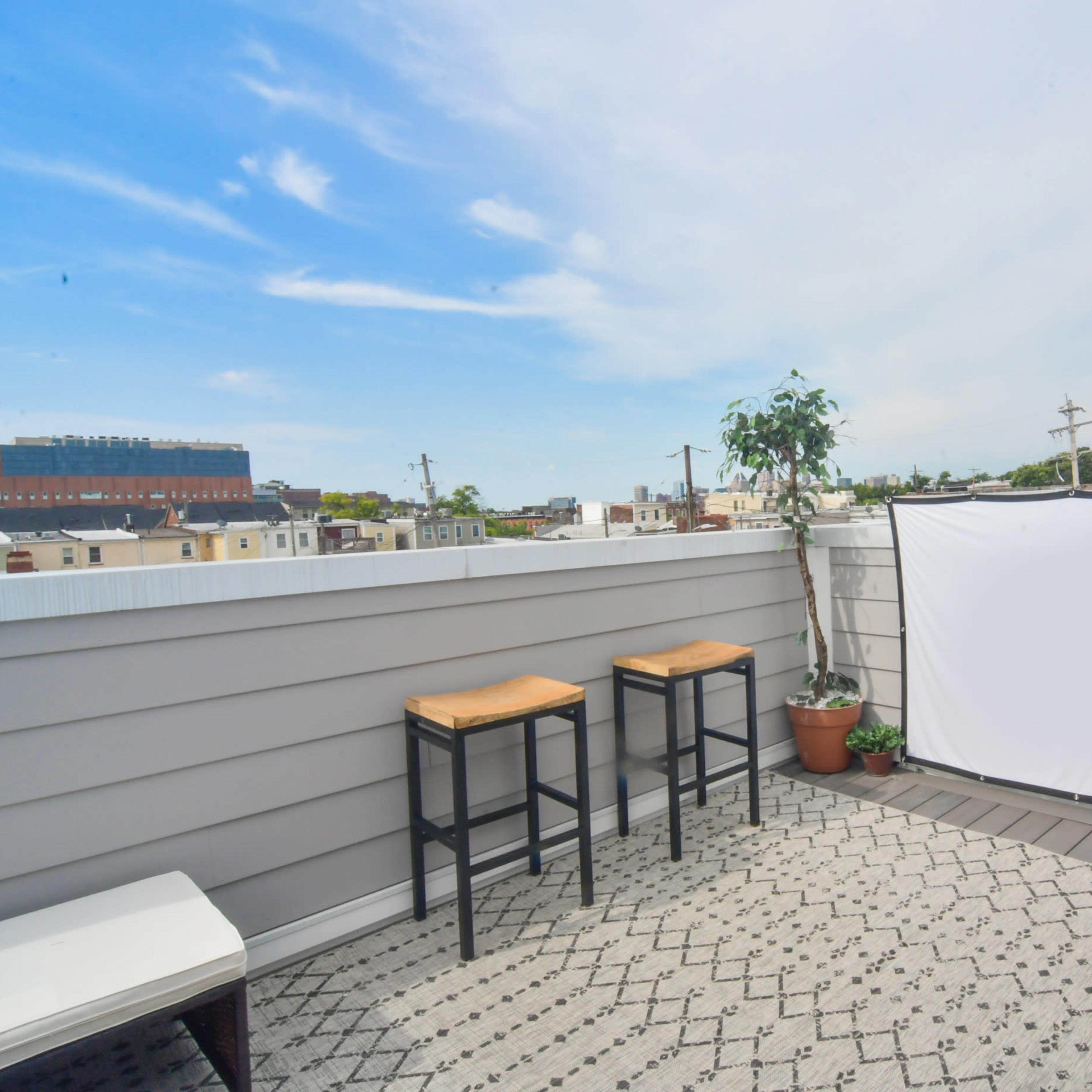 A rooftop terrace features two black chairs, a white backdrop, potted plants, and a scenic view of the skyline and church steeple.