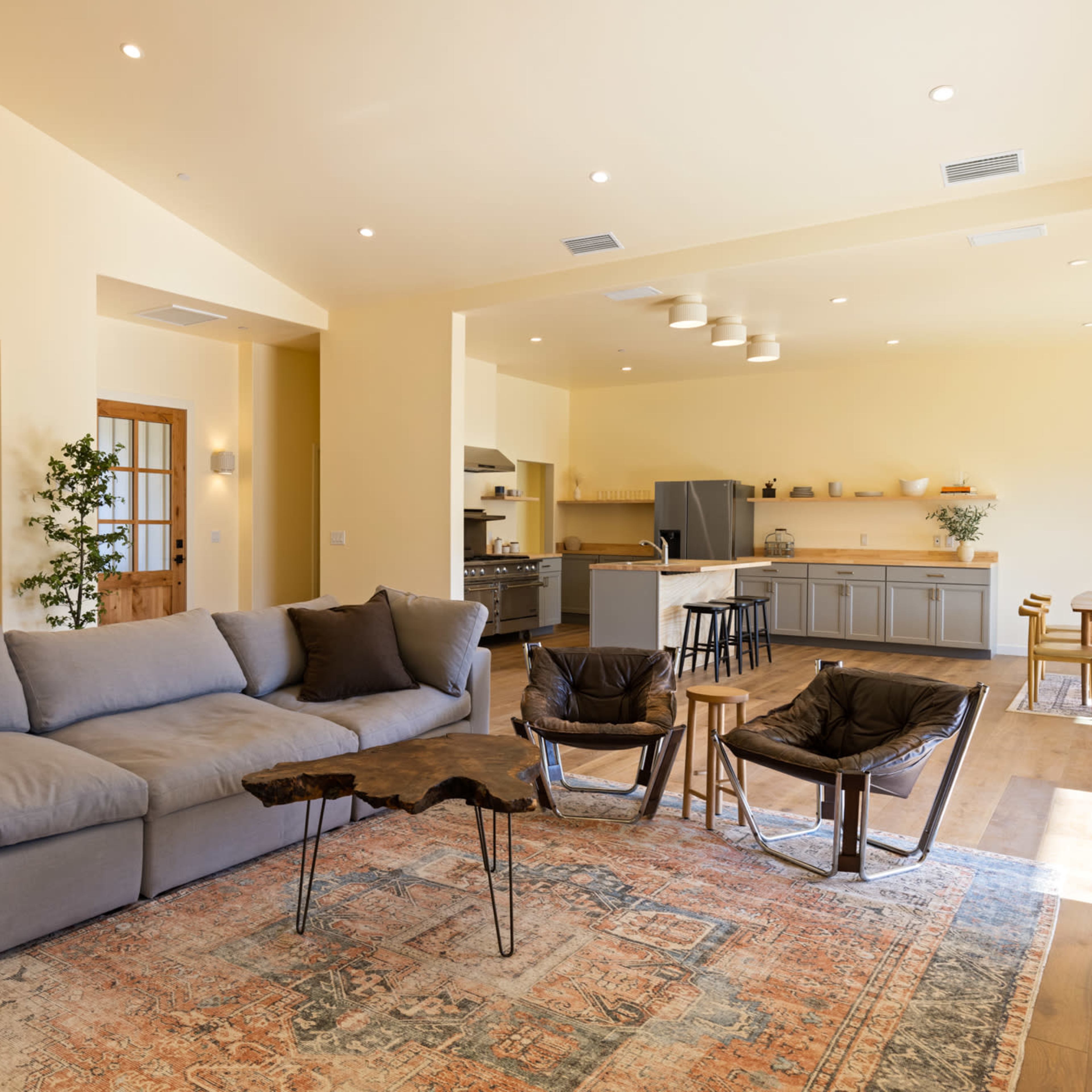 A modern living space with a gray sectional sofa, wooden coffee table, and two armchairs, leading to a kitchen area with gray cabinets and a dining table.