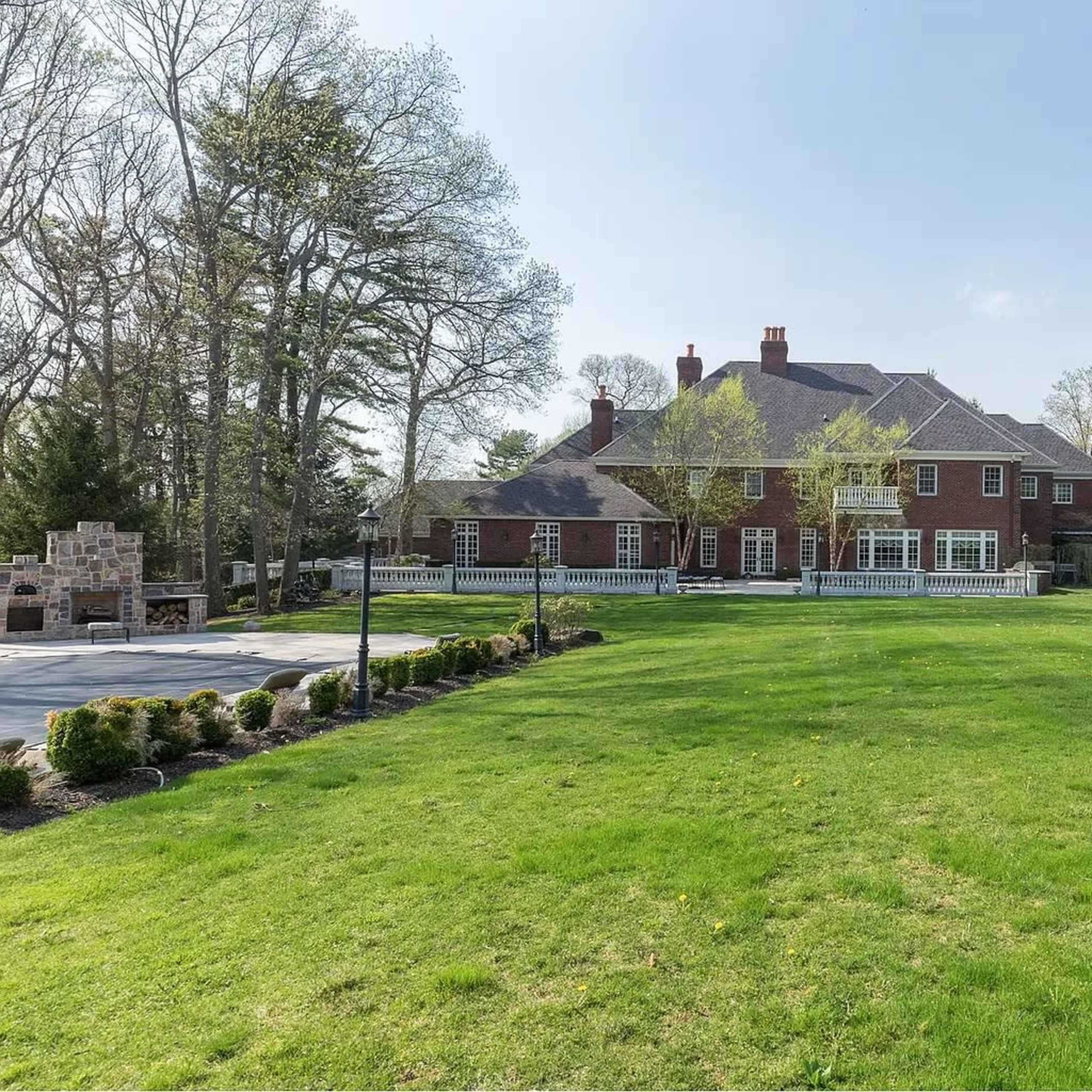 A large brick mansion is situated on a green lawn surrounded by trees and landscaped gardens.