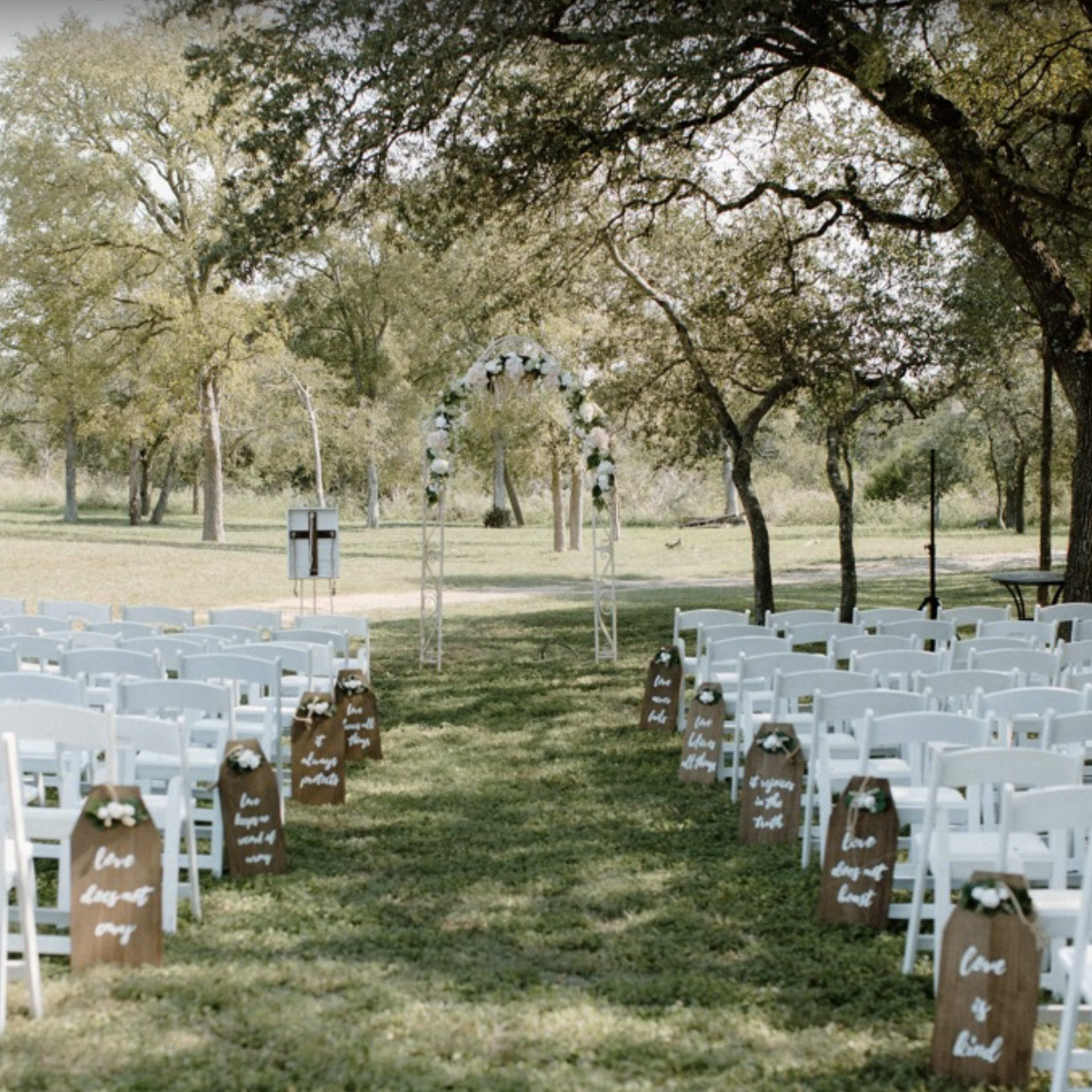 An outdoor wedding ceremony setup features rows of white chairs lining a grassy aisle leading to an arbor decorated with greenery and flowers.