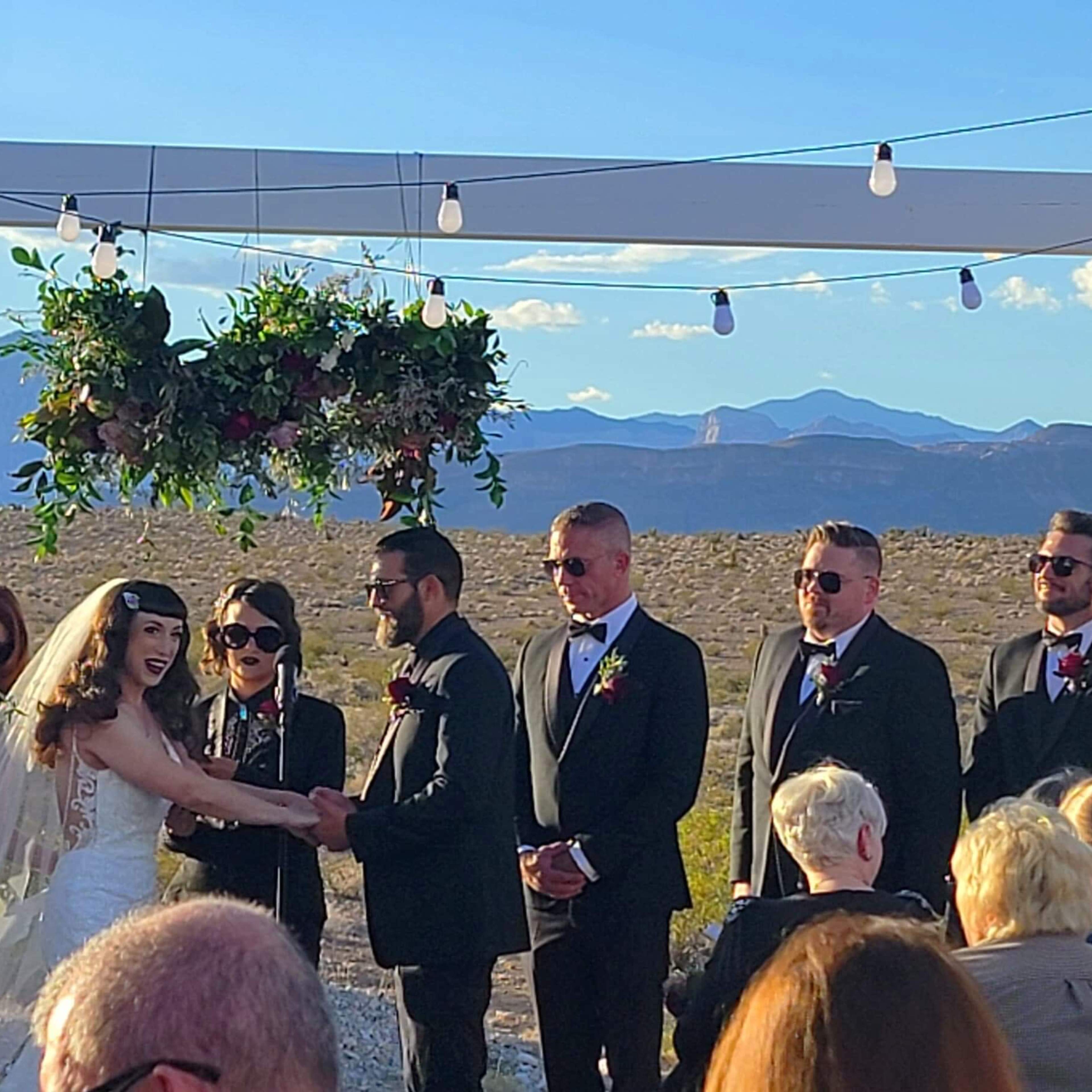 A bride and groom exchange vows under a floral arch while guests look on at an outdoor wedding ceremony in a mountainous landscape.