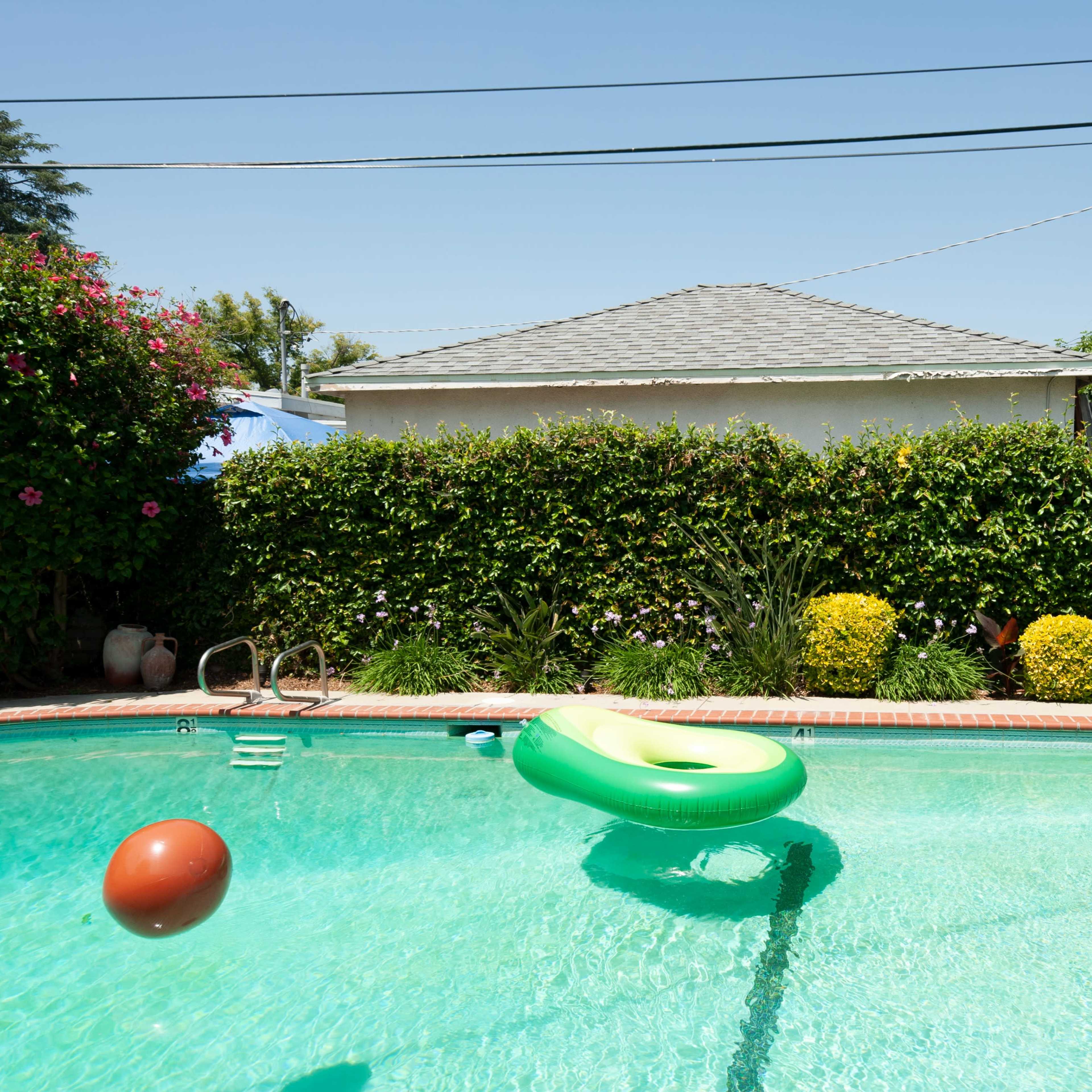 A pool with a green inflatable ring and a brown ball floats on the water, surrounded by lush greenery and a house in the background.