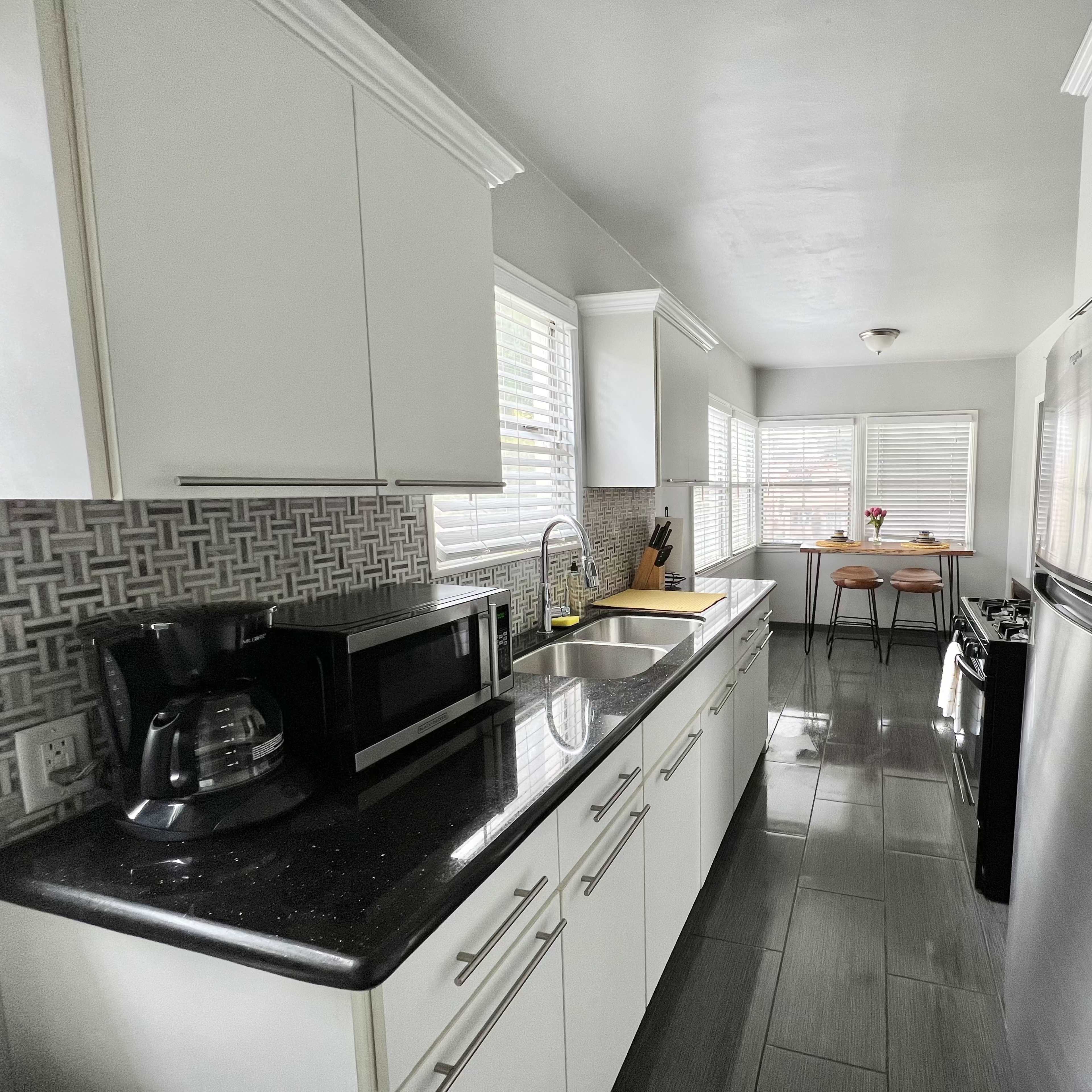 A modern kitchen features white cabinets, stainless steel appliances, and a view of a dining area with two stools and a window.