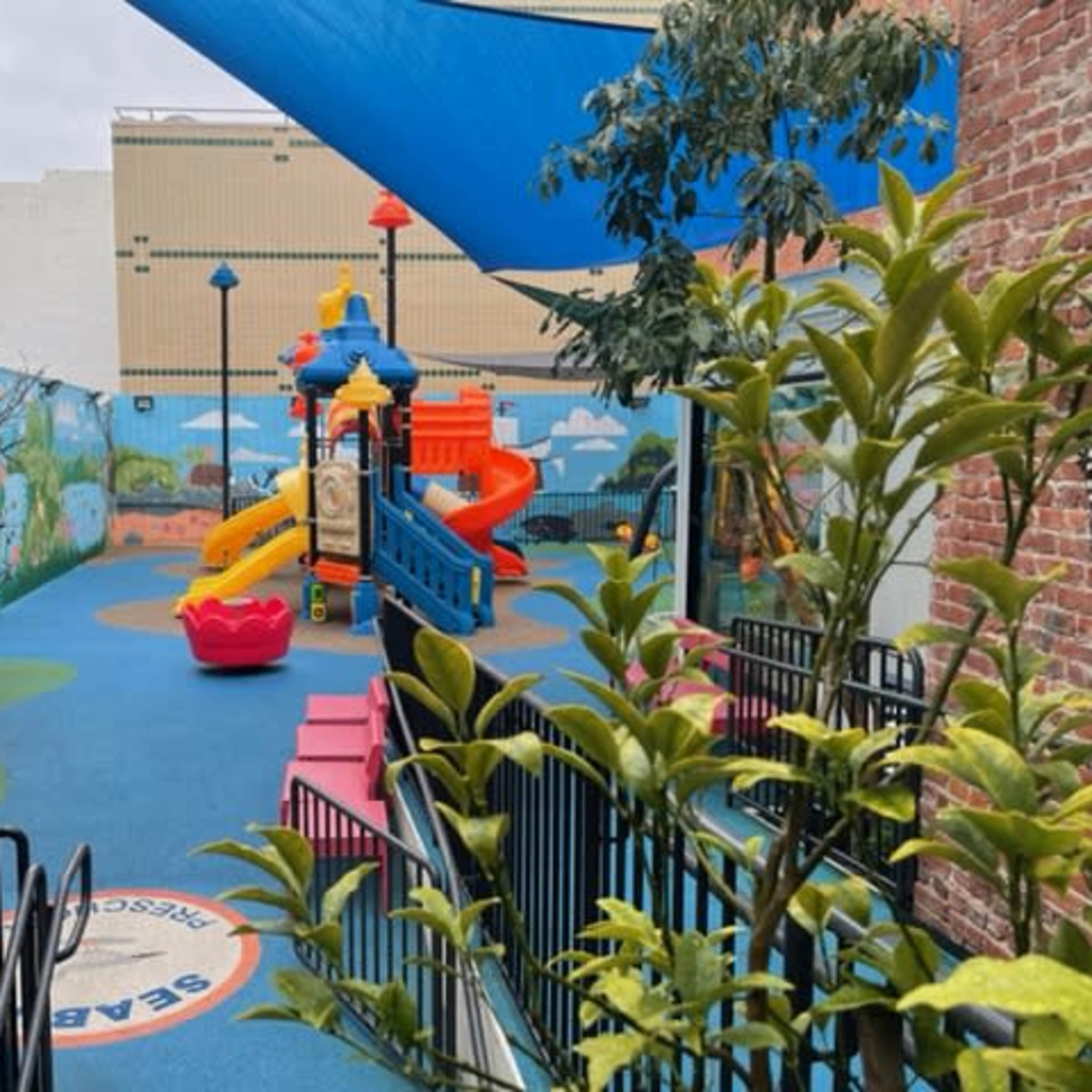 A colorful playground area with a slide and climbing structure, surrounded by greenery and mural-decorated walls.
