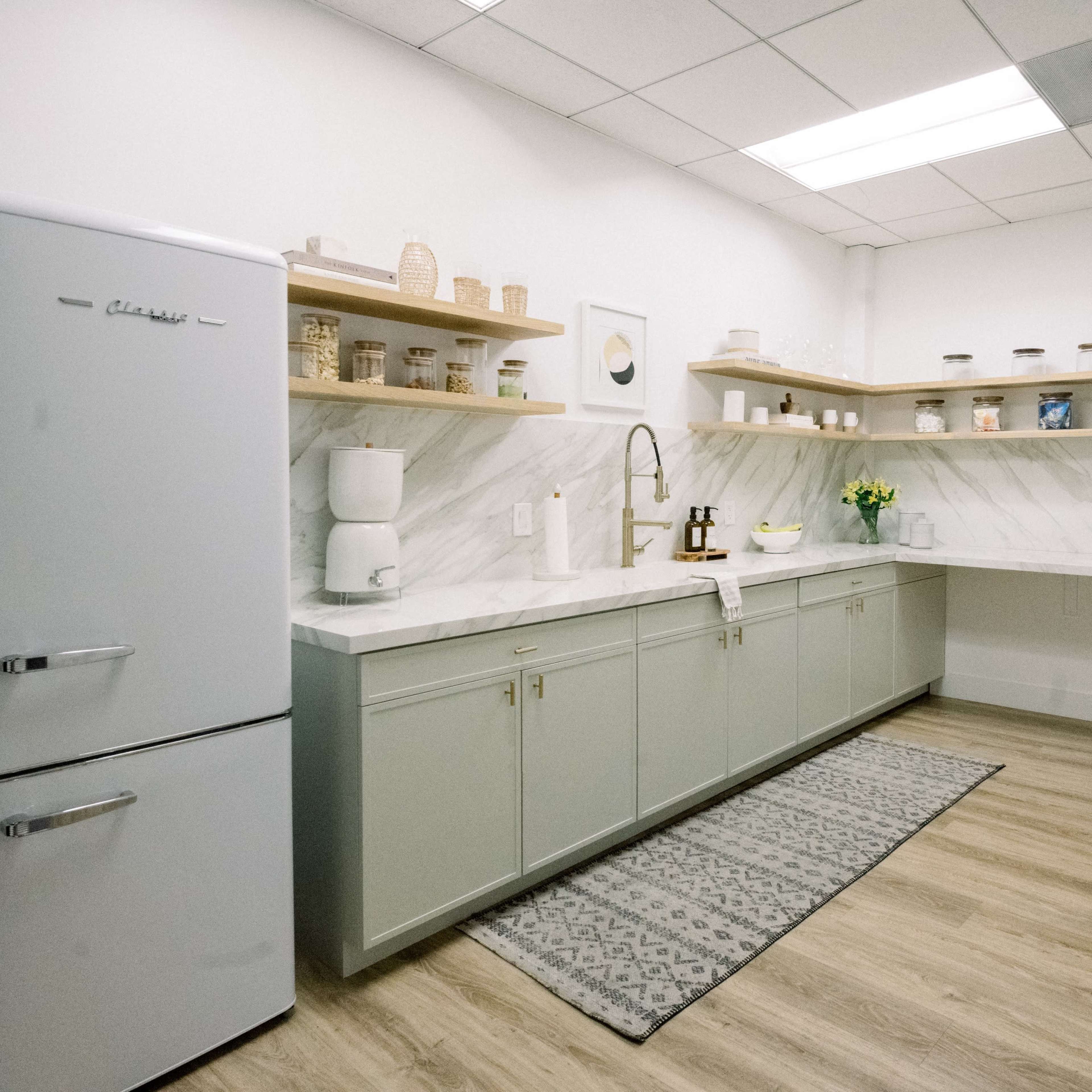 A modern kitchen features light green cabinets, a white marble countertop, and neatly arranged shelves holding jars and decorative items.