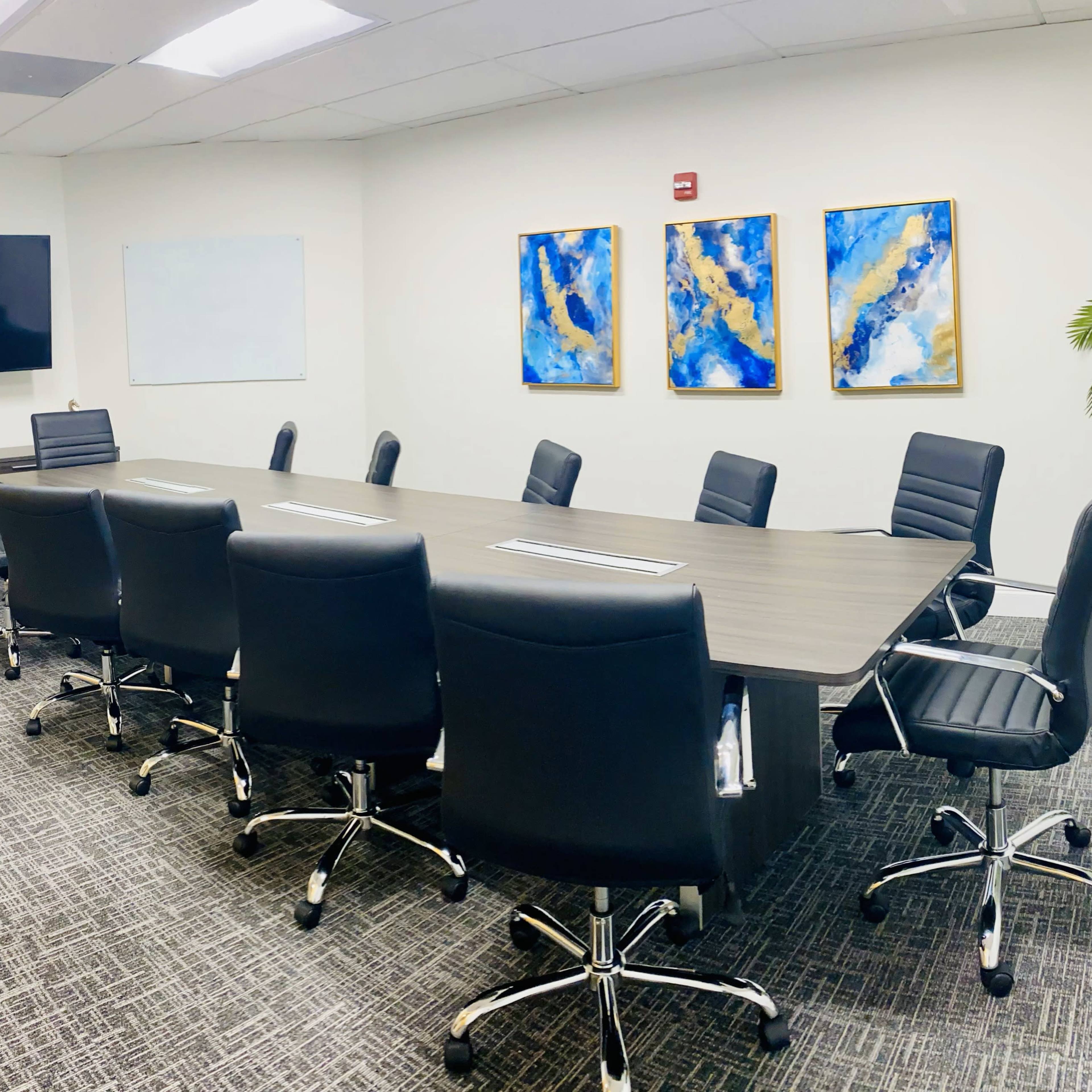 A modern conference room features a long table surrounded by black leather chairs, with a television, whiteboard, and abstract art on the walls.