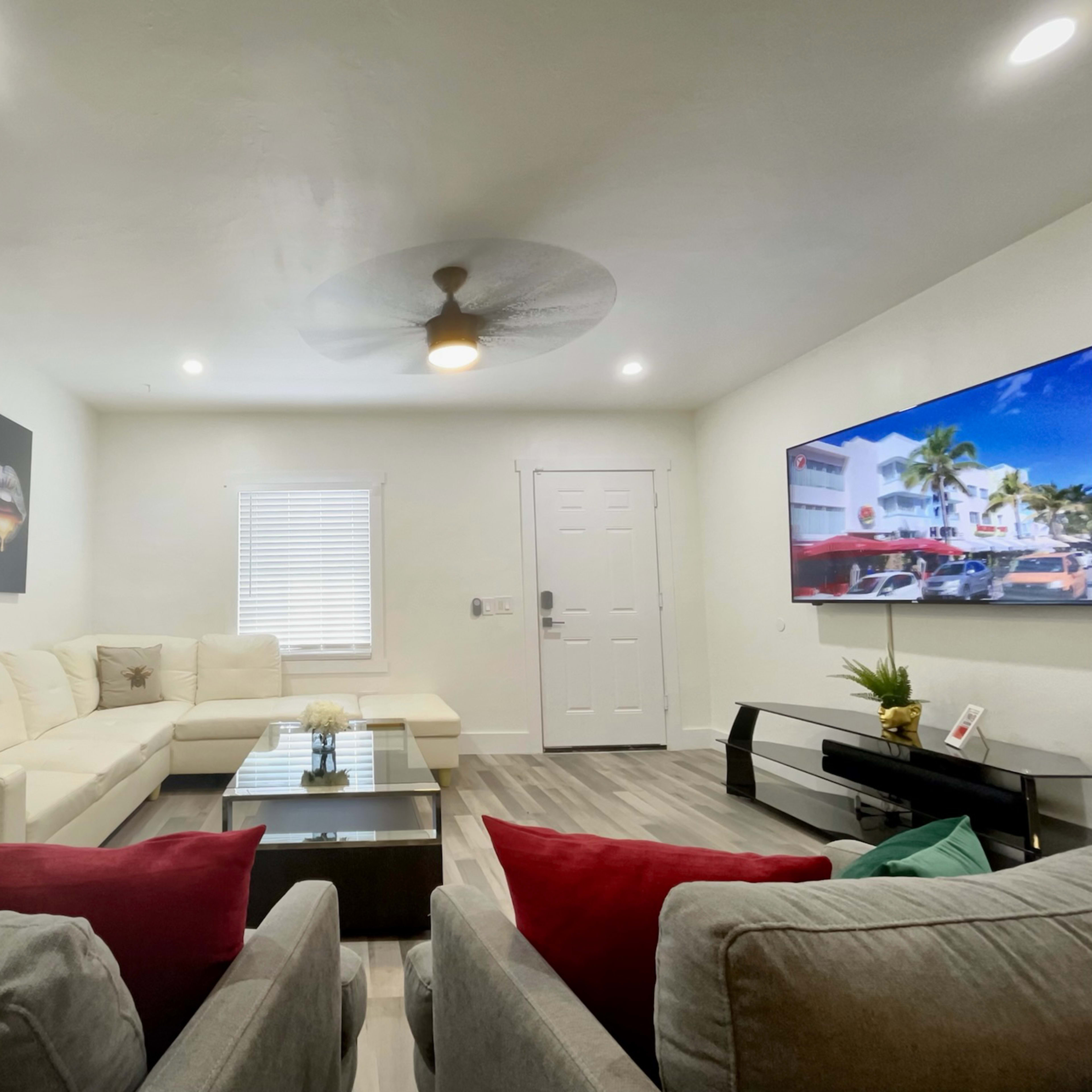 A modern living room with a white sectional sofa, two gray chairs with red and green cushions, a coffee table, wall art, and a large flat-screen TV.