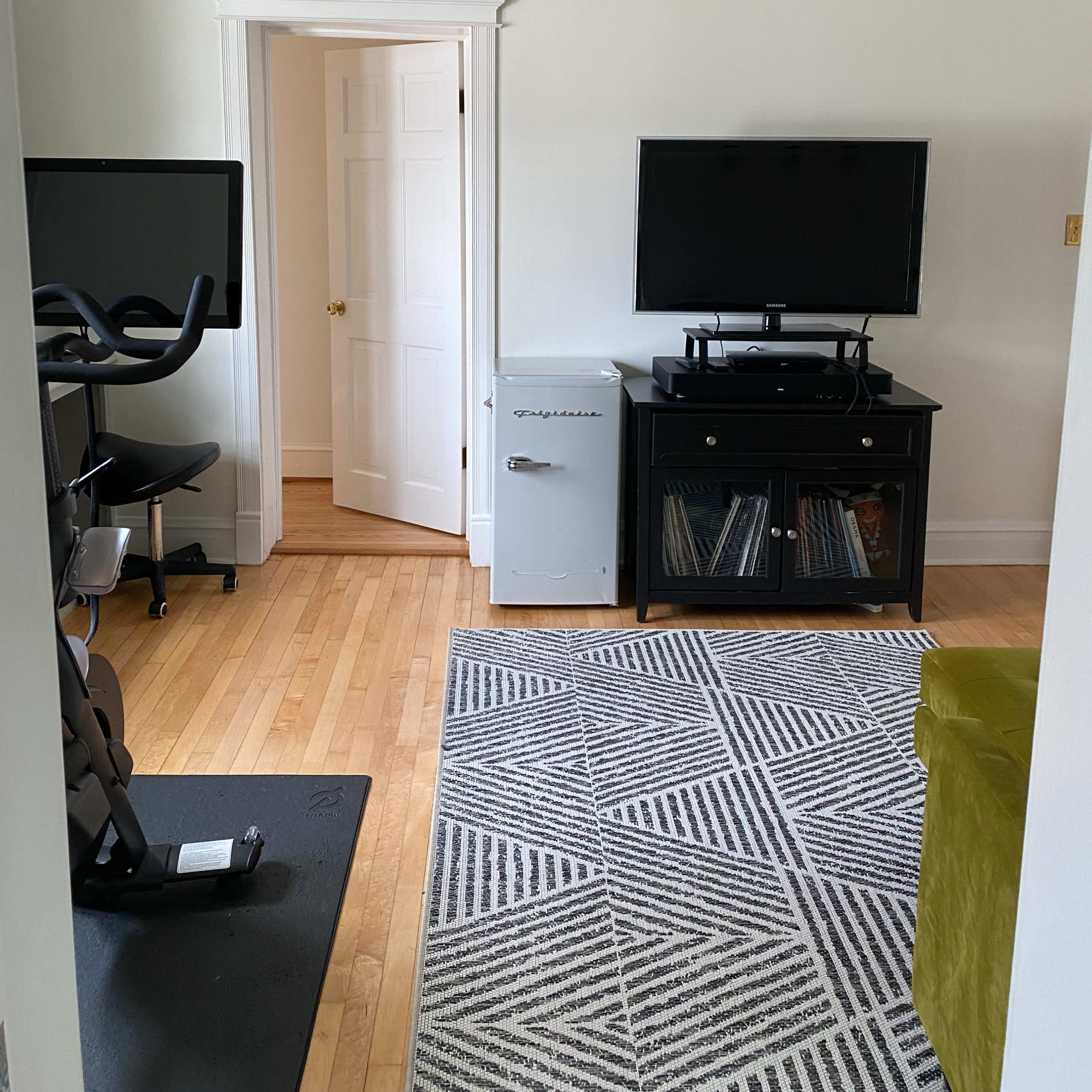A small room with a bike, a television on a cabinet, and a mini fridge, with a door leading to another space.