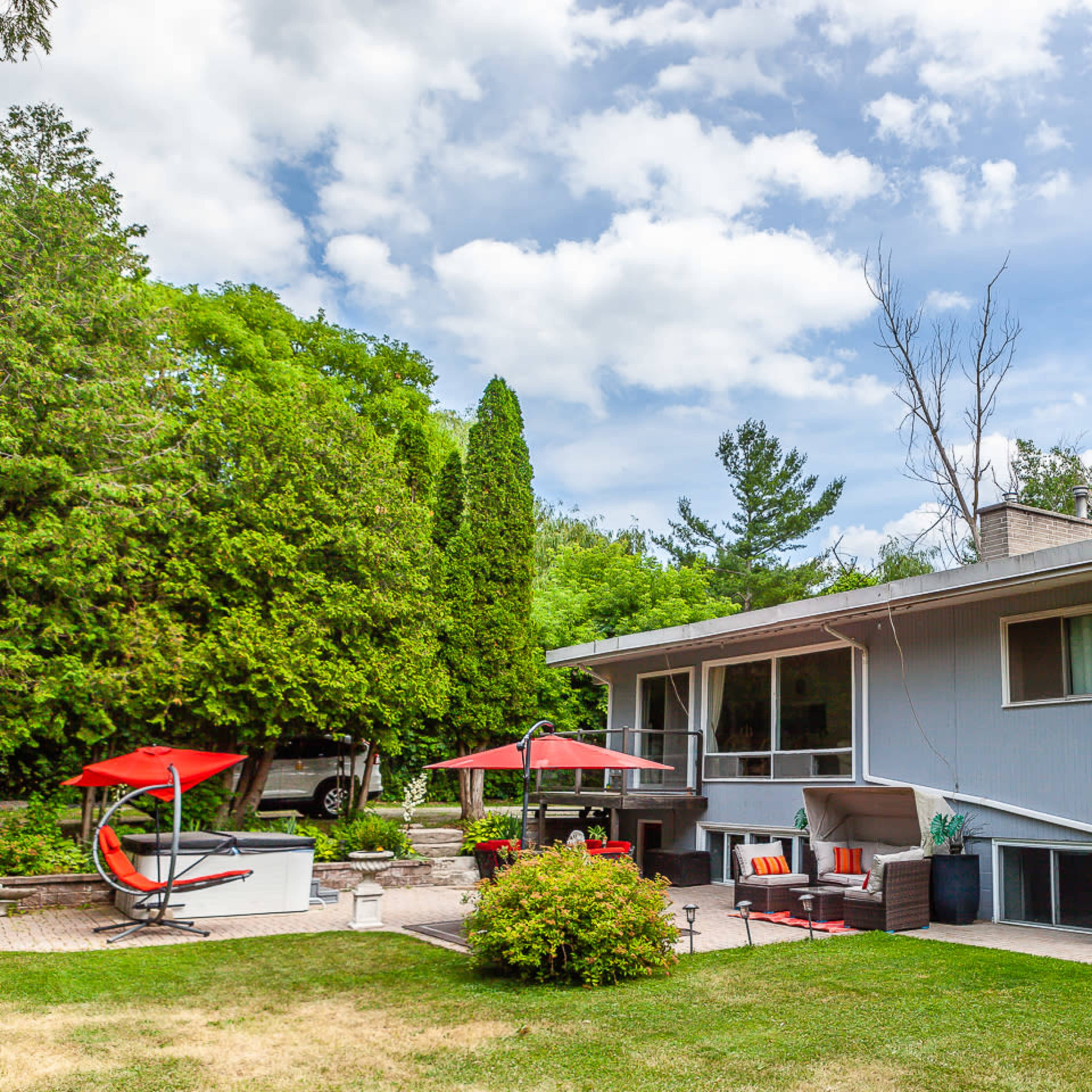 A light blue house with a landscaped yard features outdoor seating areas and a patio under red umbrellas.