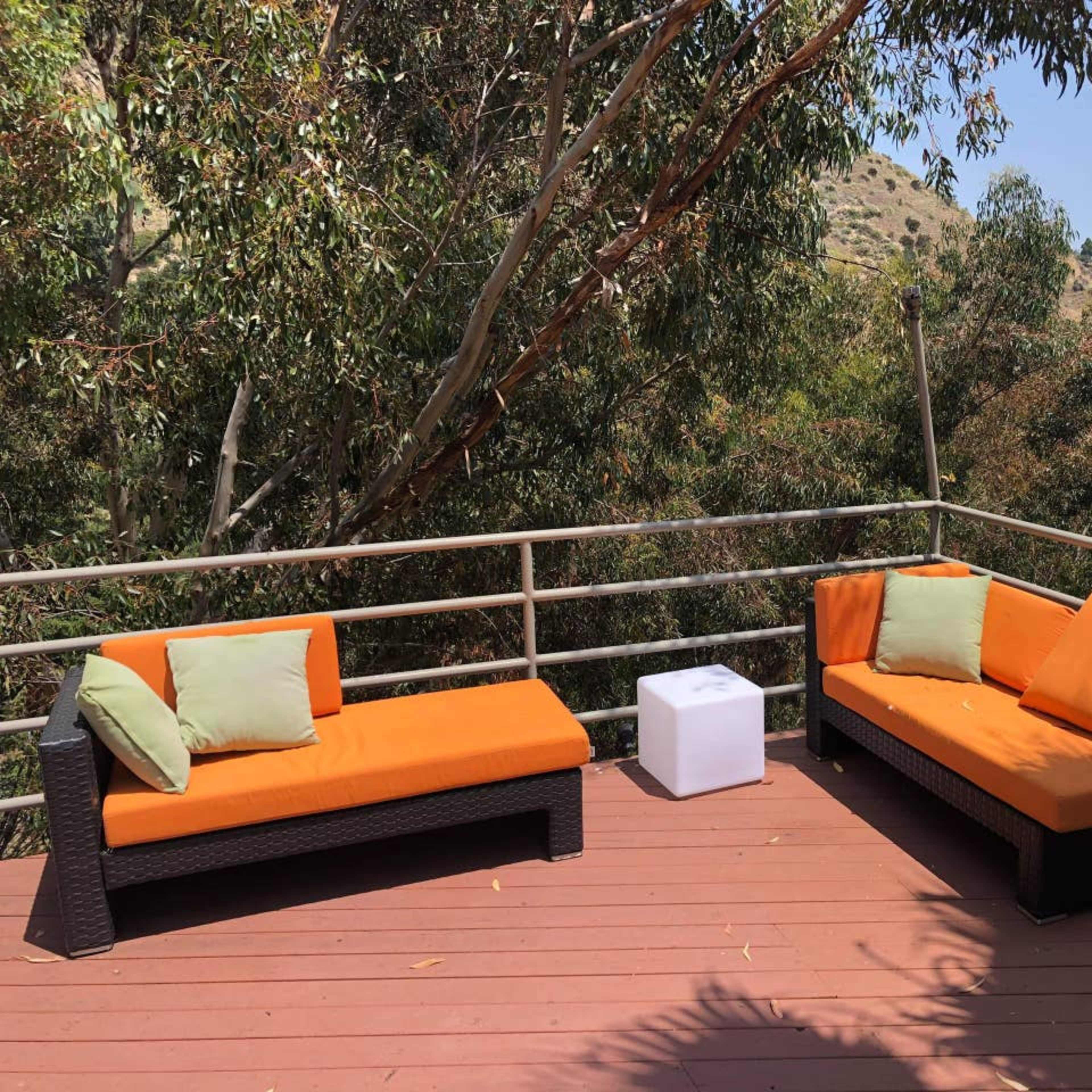 A deck with two orange couches and white cube tables, surrounded by tall green trees.