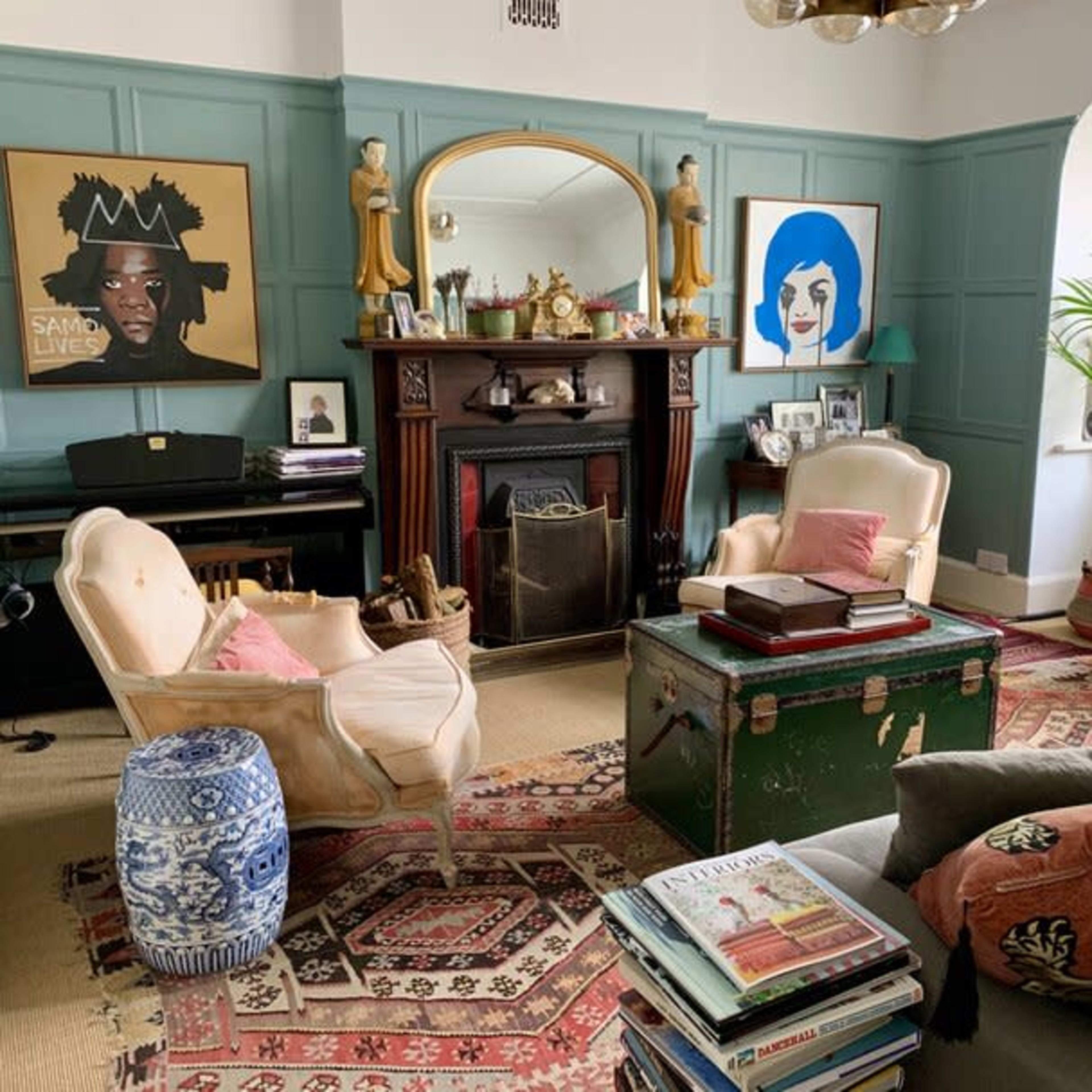 A cozy living room features two armchairs, a green coffee table, a vintage-style fireplace, and a mix of artwork on the walls.