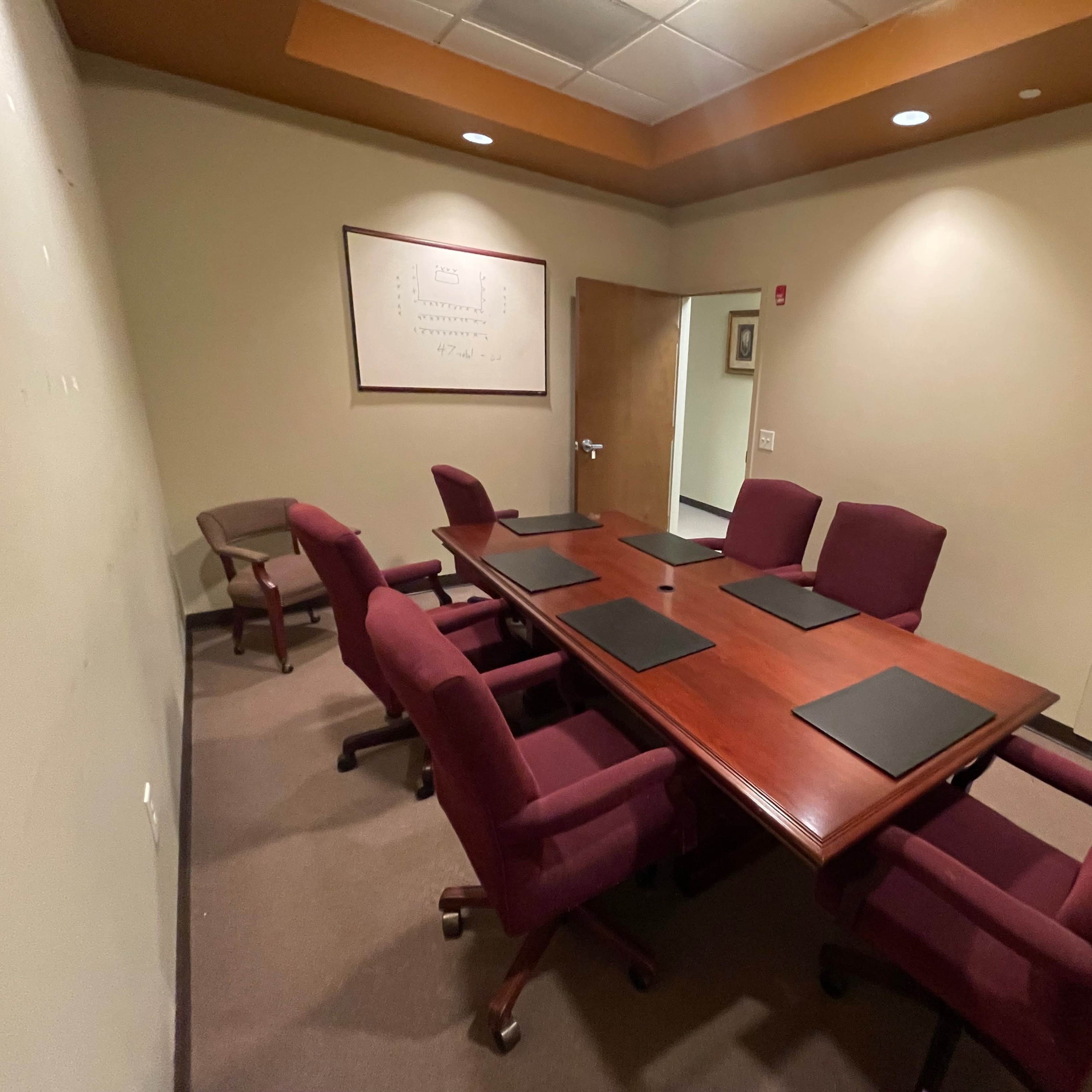 A conference room features a large wooden table surrounded by six maroon chairs, with a whiteboard and a closed door visible in the background.