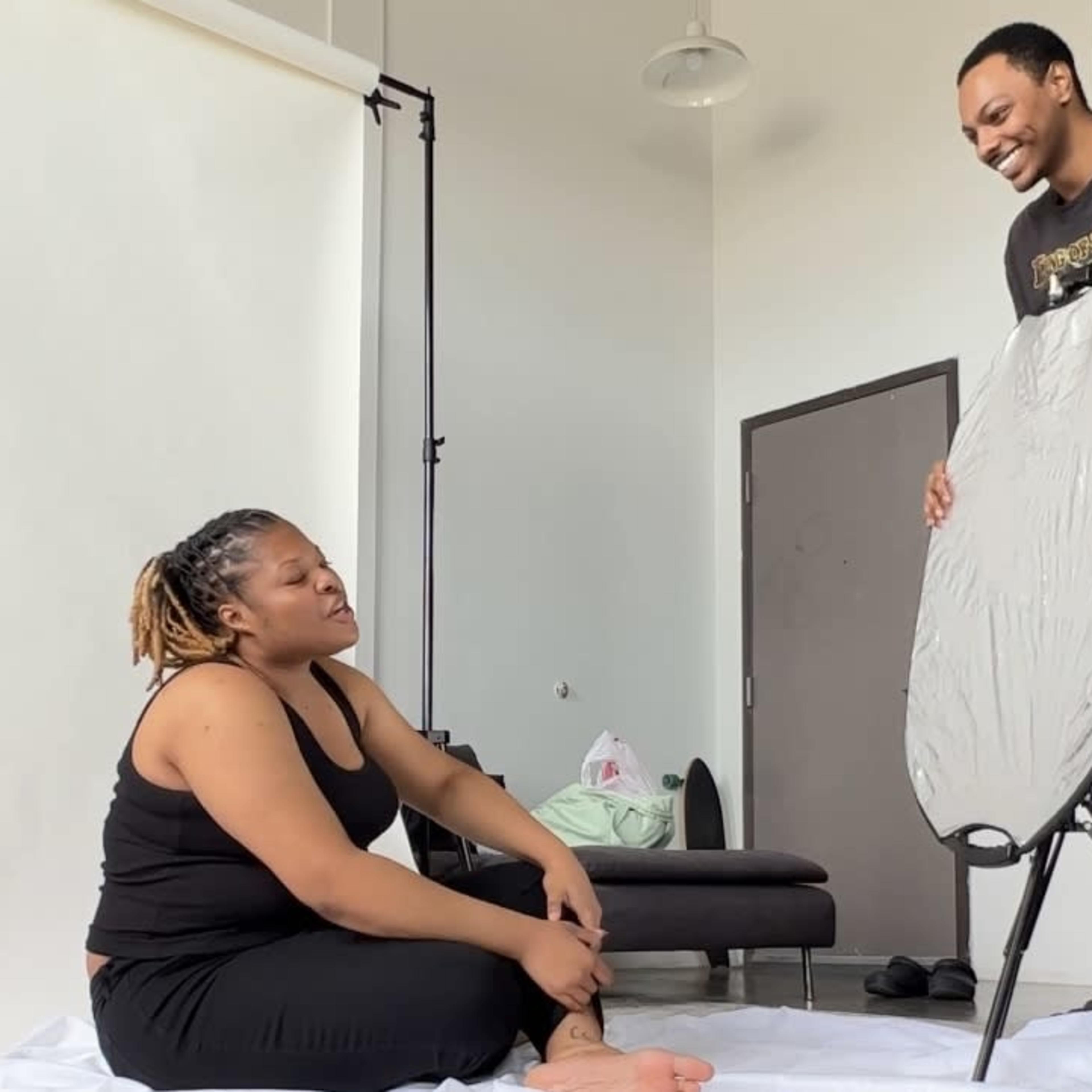 A woman sits on a white backdrop while a man sets up an ironing board in a well-lit room.