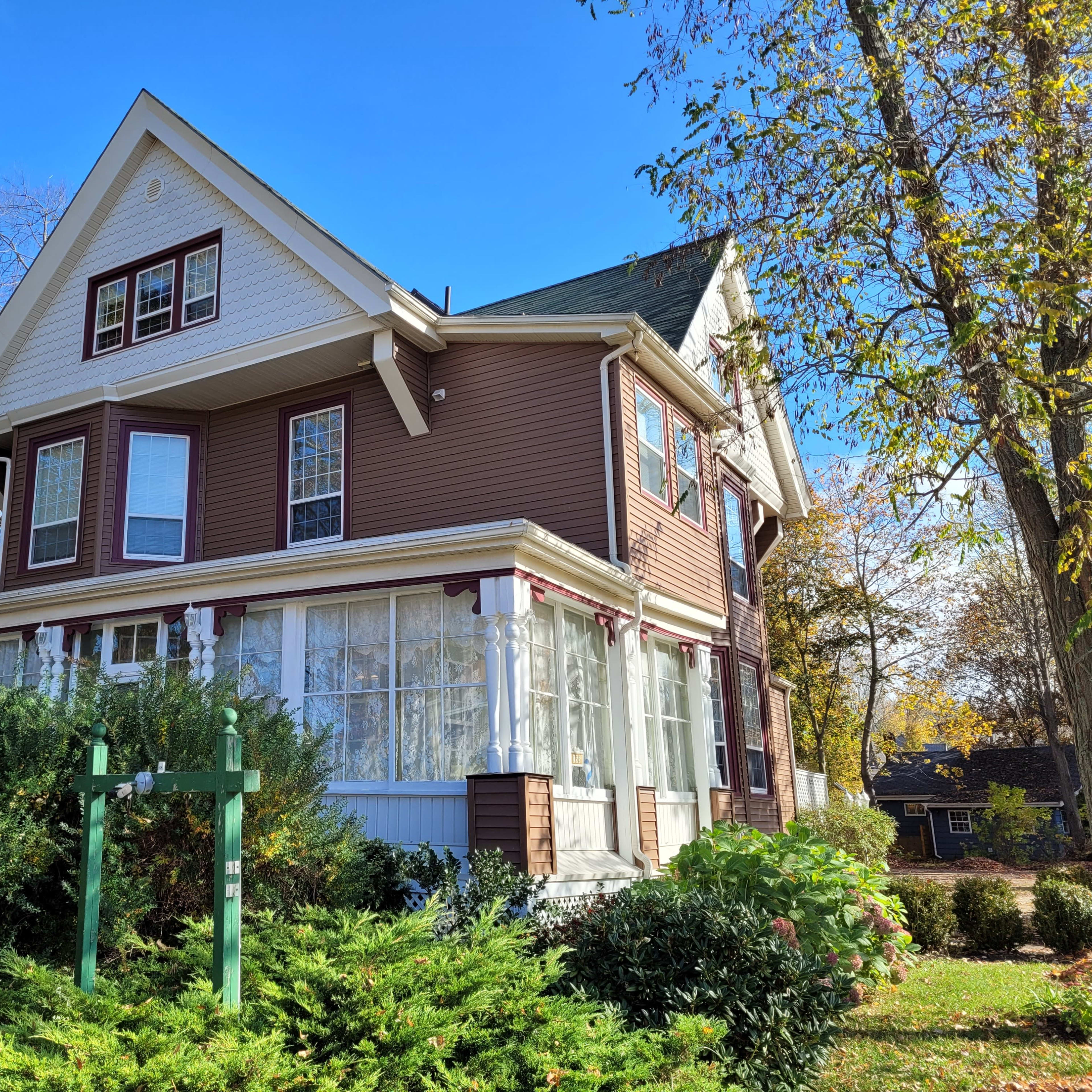 A large Victorian-style house with a wraparound porch is surrounded by greenery and trees.