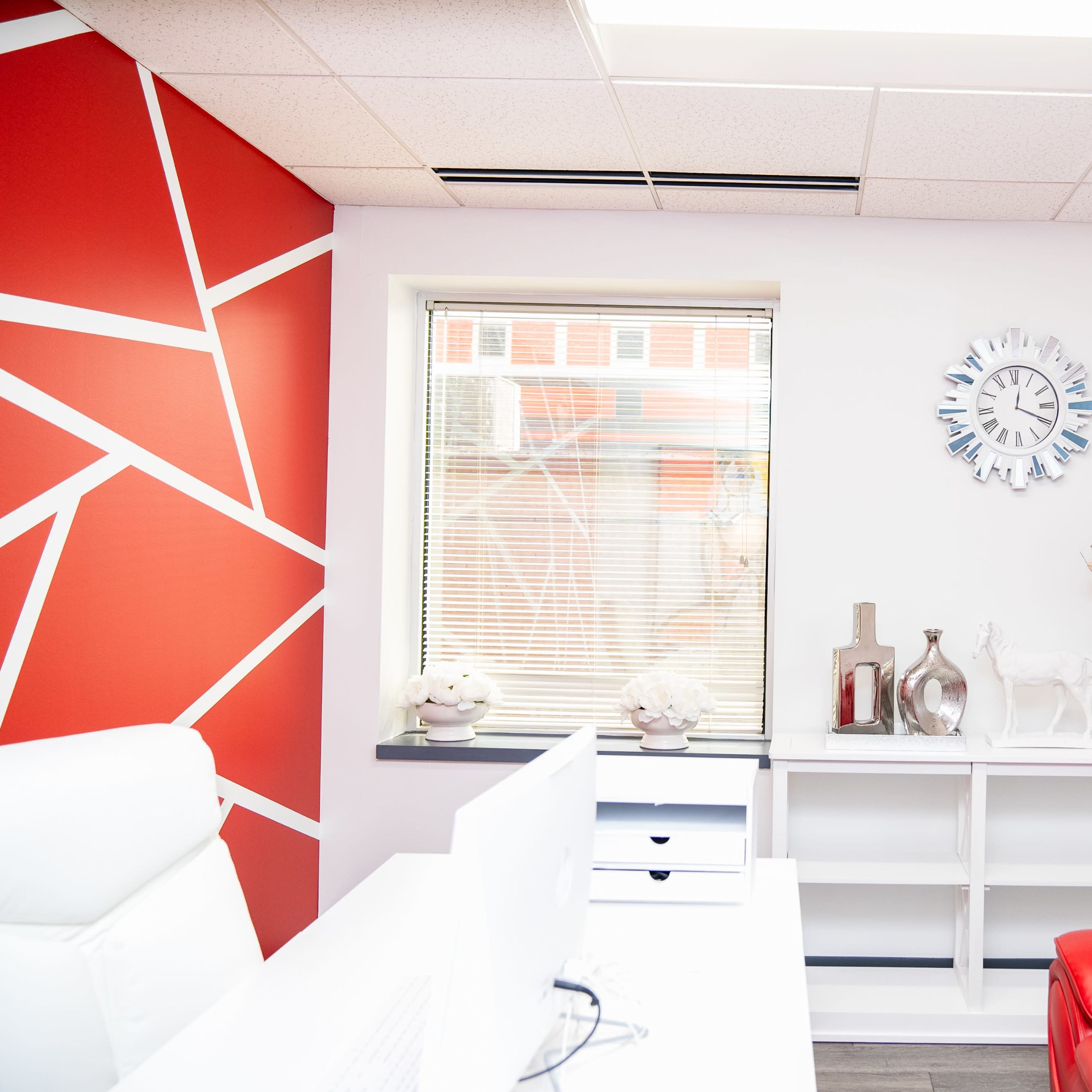 A modern office space with a red accent wall, a white desk with a computer, and a red couch beside a decorative shelf.