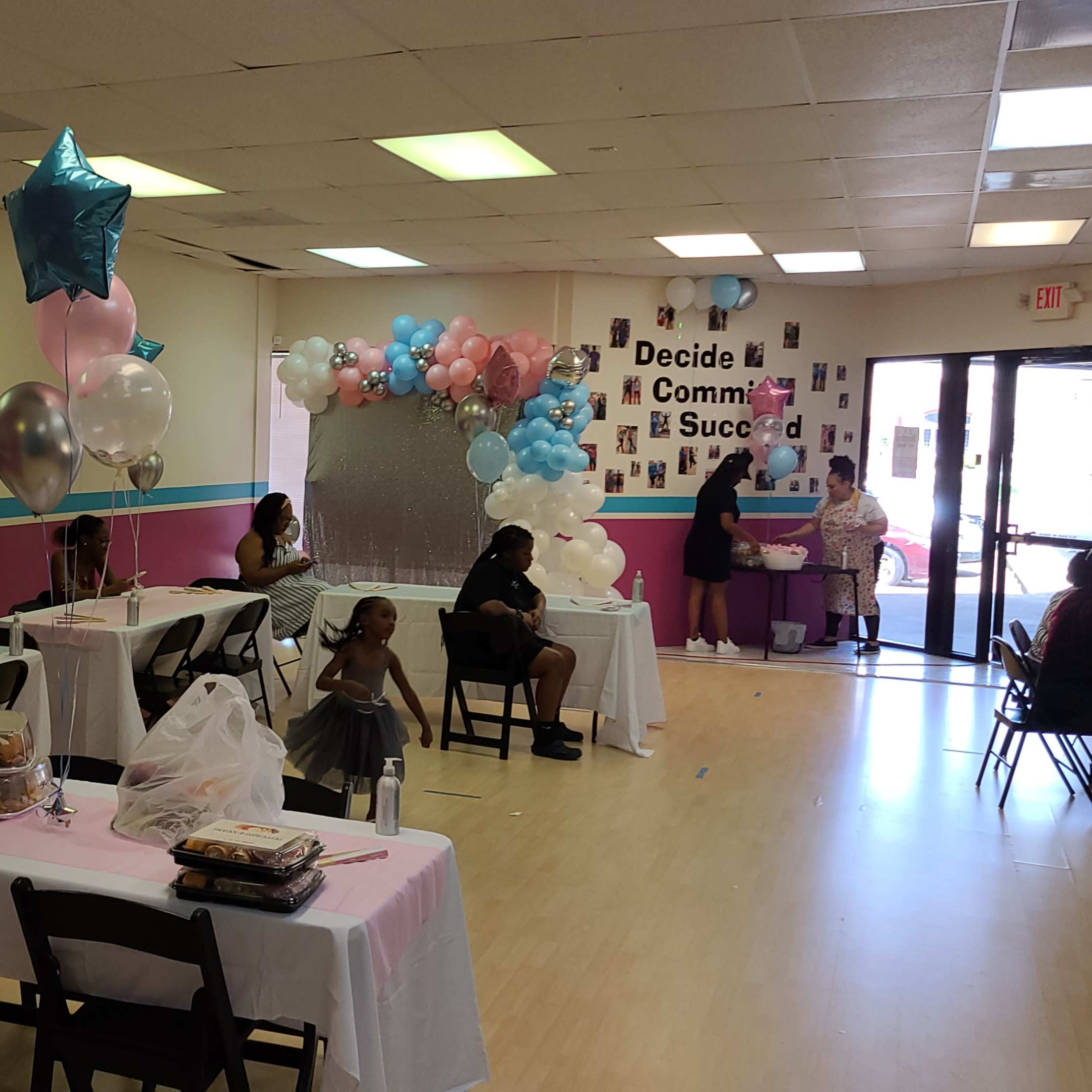A decorated event space with tables set up, balloons, and attendees engaged in activities.
