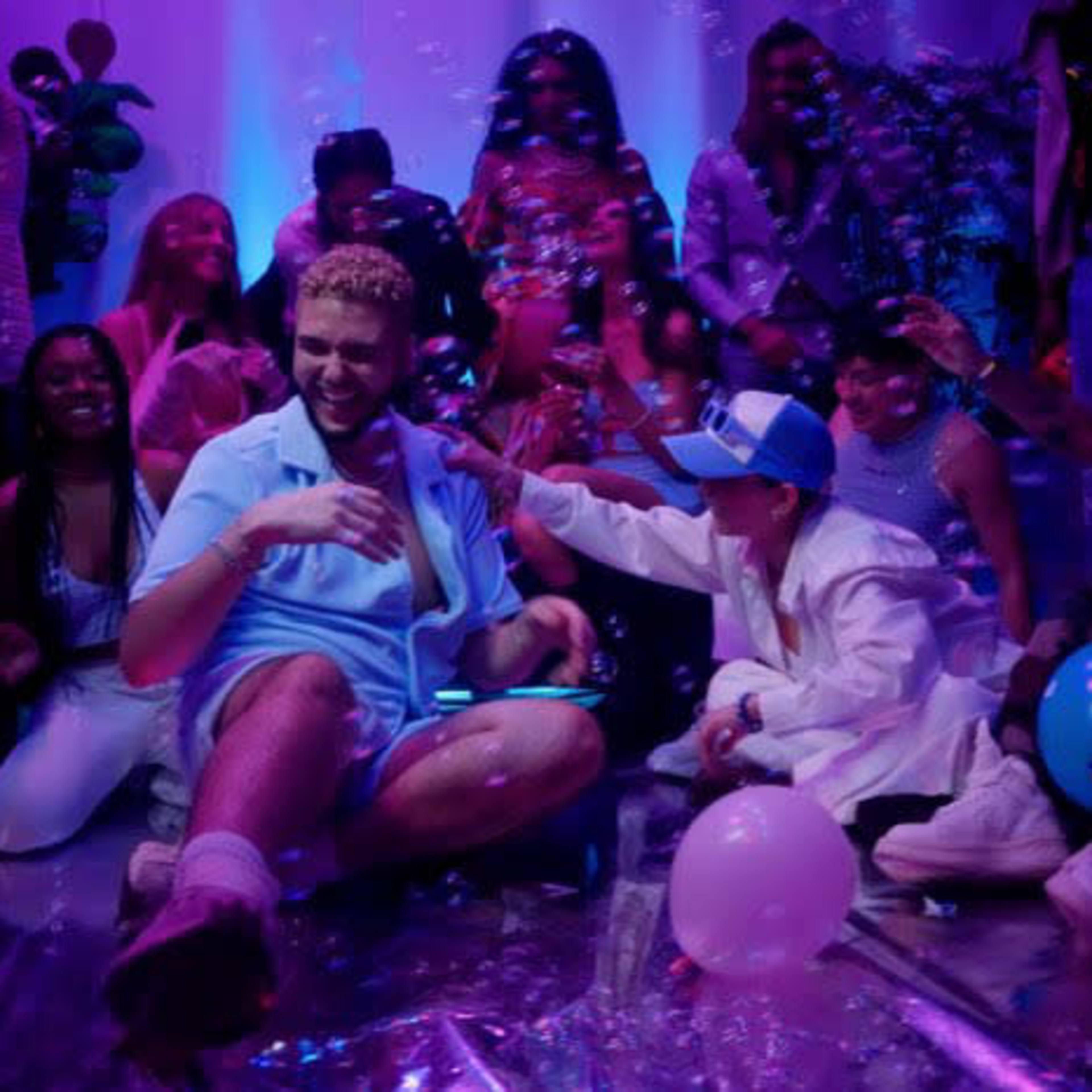 A group of people in colorful attire celebrate at a lively party, surrounded by bubbles and balloons, with one person sitting prominently on the floor.